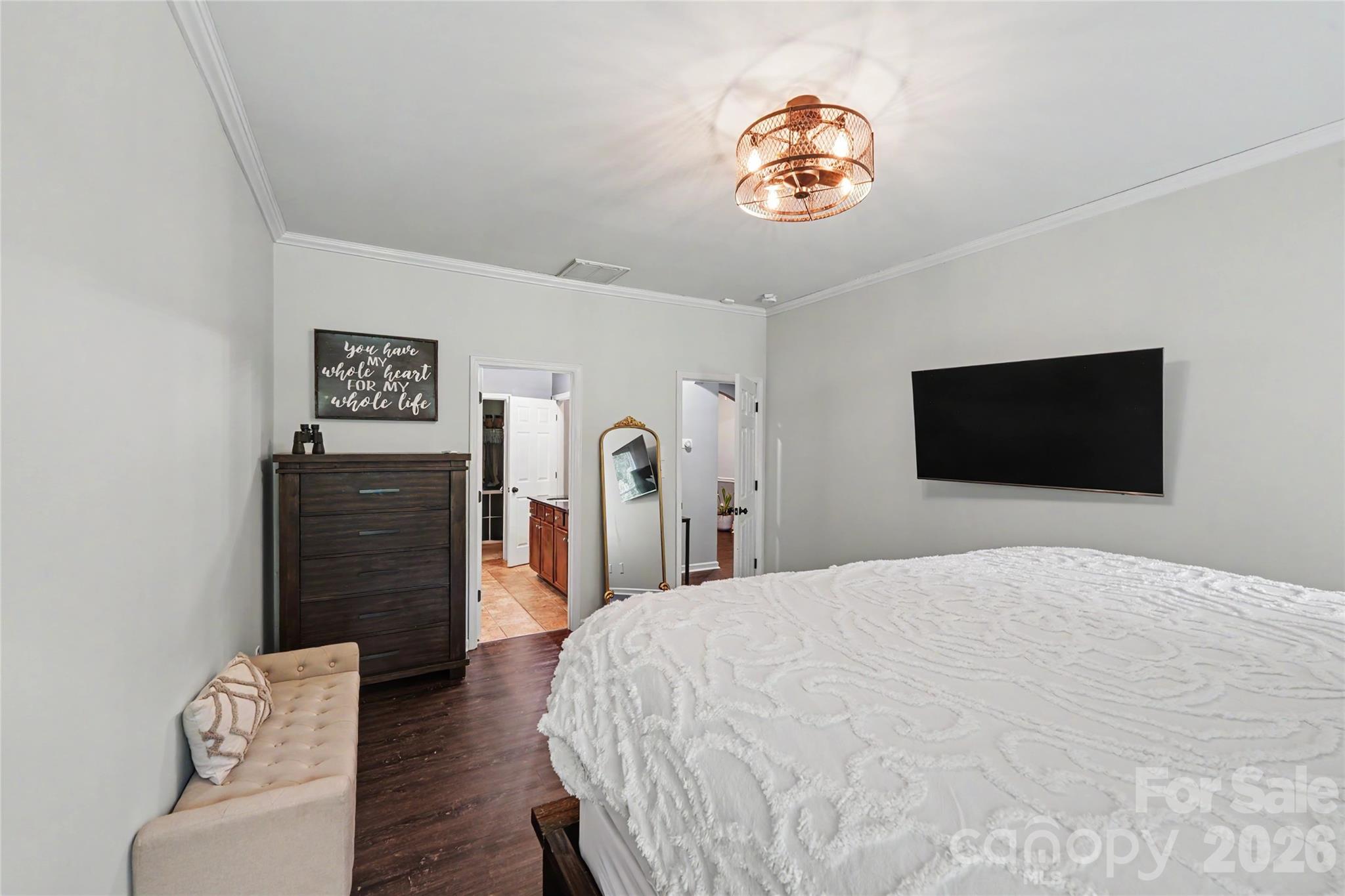 Primary bedroom with a white textured bedspread, wall-mounted TV, dark dresser, and adjoining doorways.
