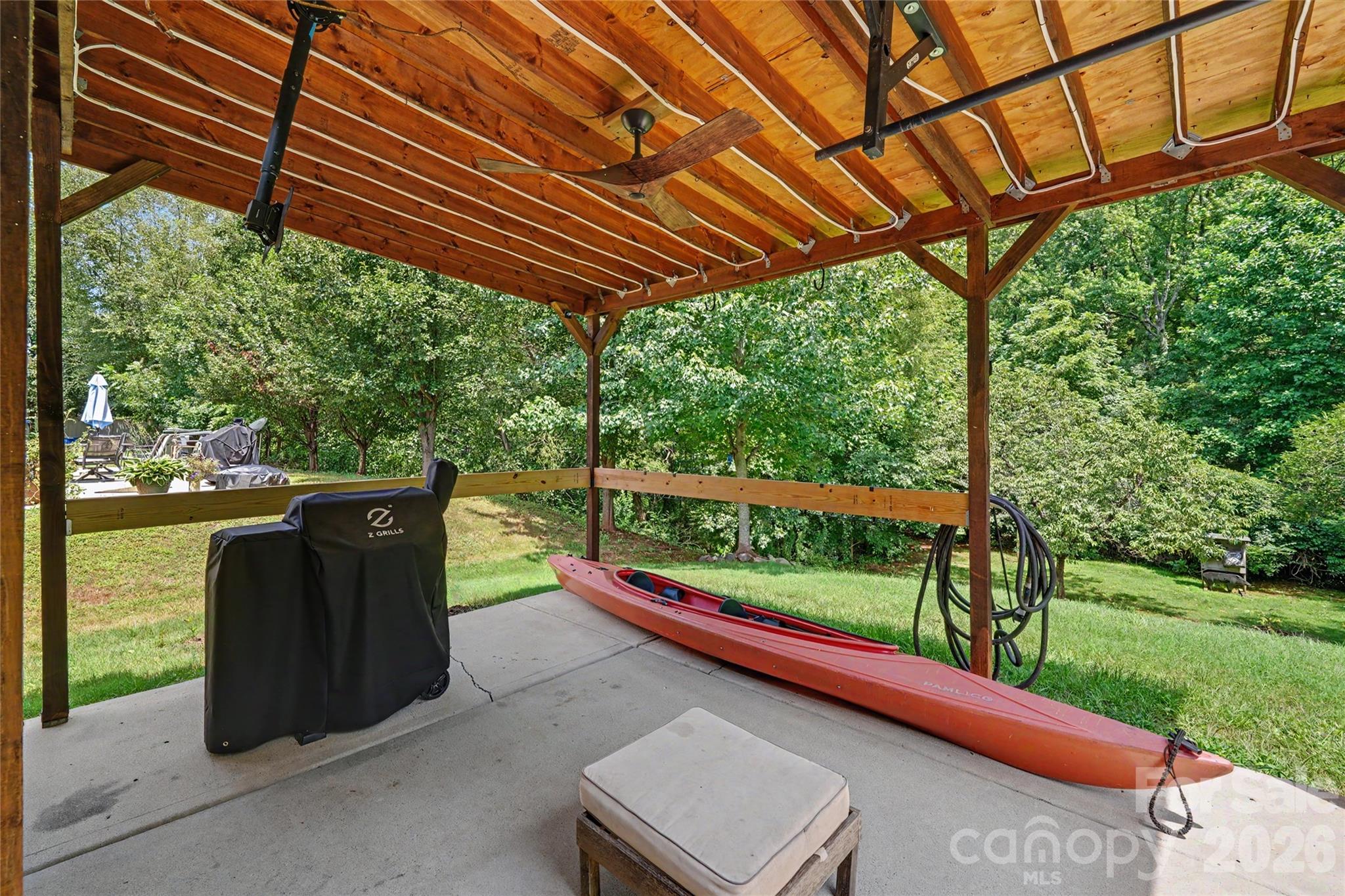 Covered timber patio with a grill and red kayak overlooking a tree-lined backyard.