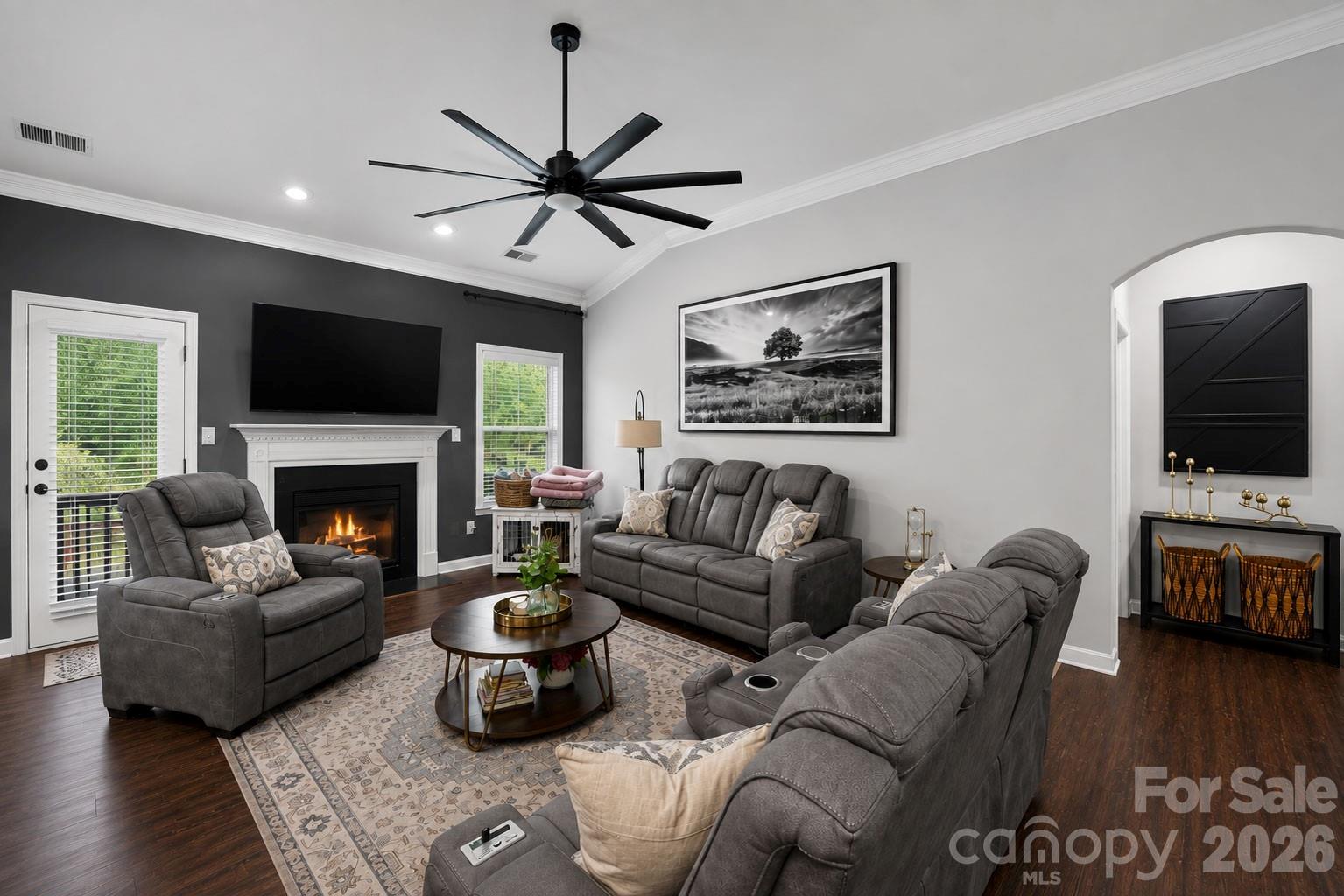Living room with gray reclining seating, a lit fireplace, and a large black ceiling fan.