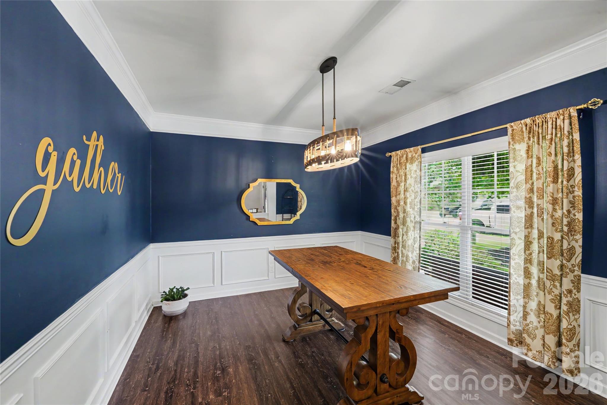Dining room with navy walls, white wainscoting, a wooden table, and gold “Gather” wall lettering.