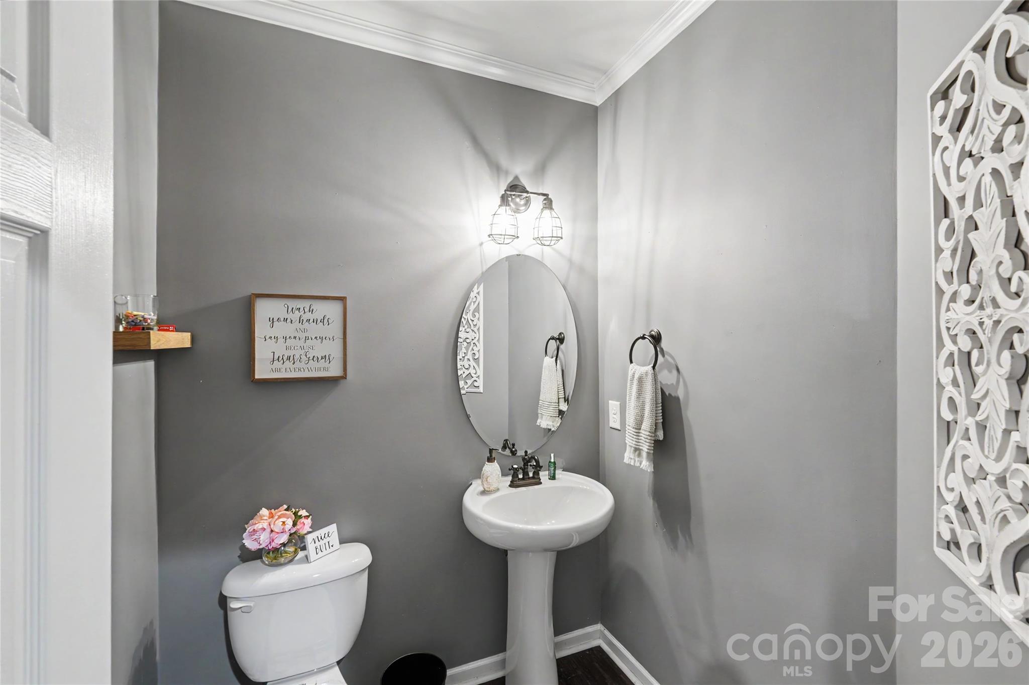 Gray bathroom with a pedestal sink, oval mirror, toilet, and decorative white wall panel.
