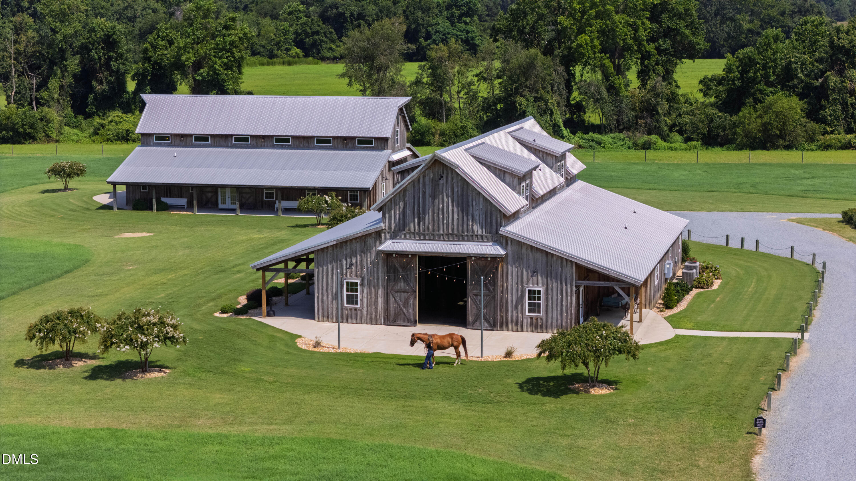 Large weathered barn with silver roofs on a green lawn, with a horse and handler by the entrance.