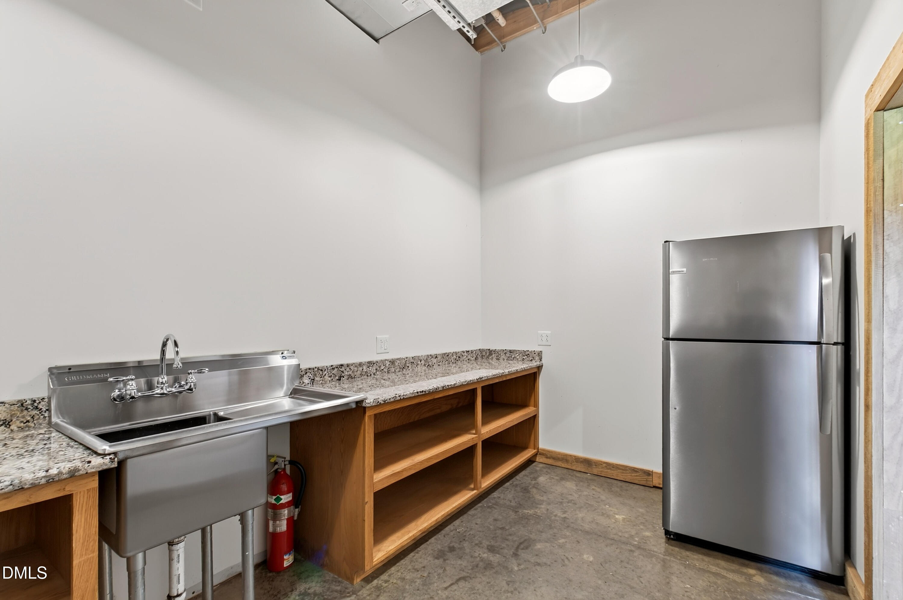 Kitchenette with a deep stainless-steel sink, granite counters, open wooden shelving, and a stainless refrigerator.