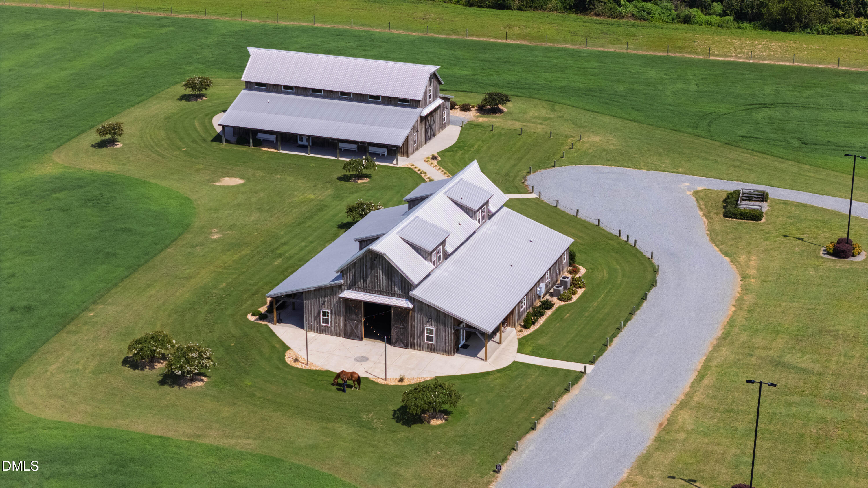Aerial view of two barn-style buildings with metal roofs, a curved gravel drive, and a grazing horse.