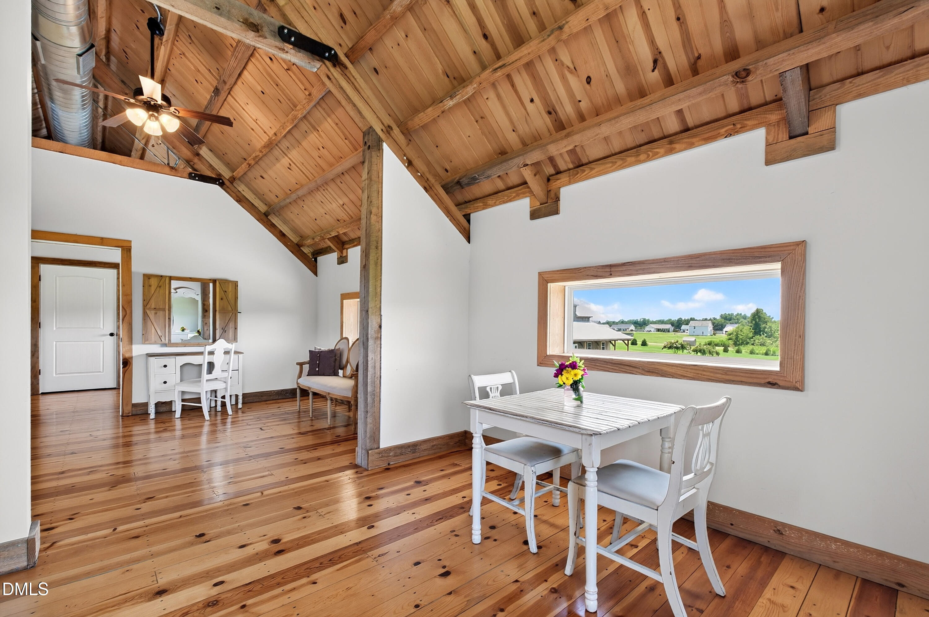 Dining area with a white table beneath an exposed timber ceiling and a wide window overlooking green lawns.
