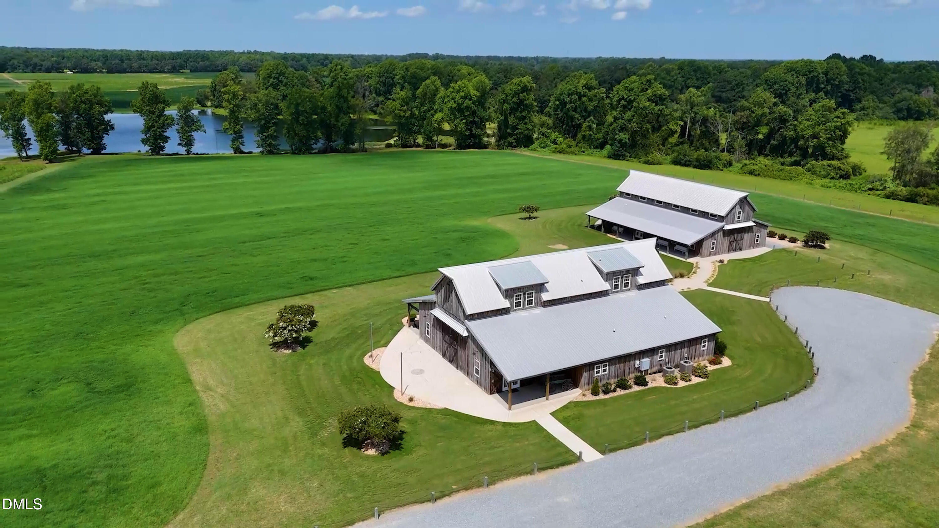 Aerial view of two barn-style buildings amid expansive lawns, with a pond and wooded tree line.