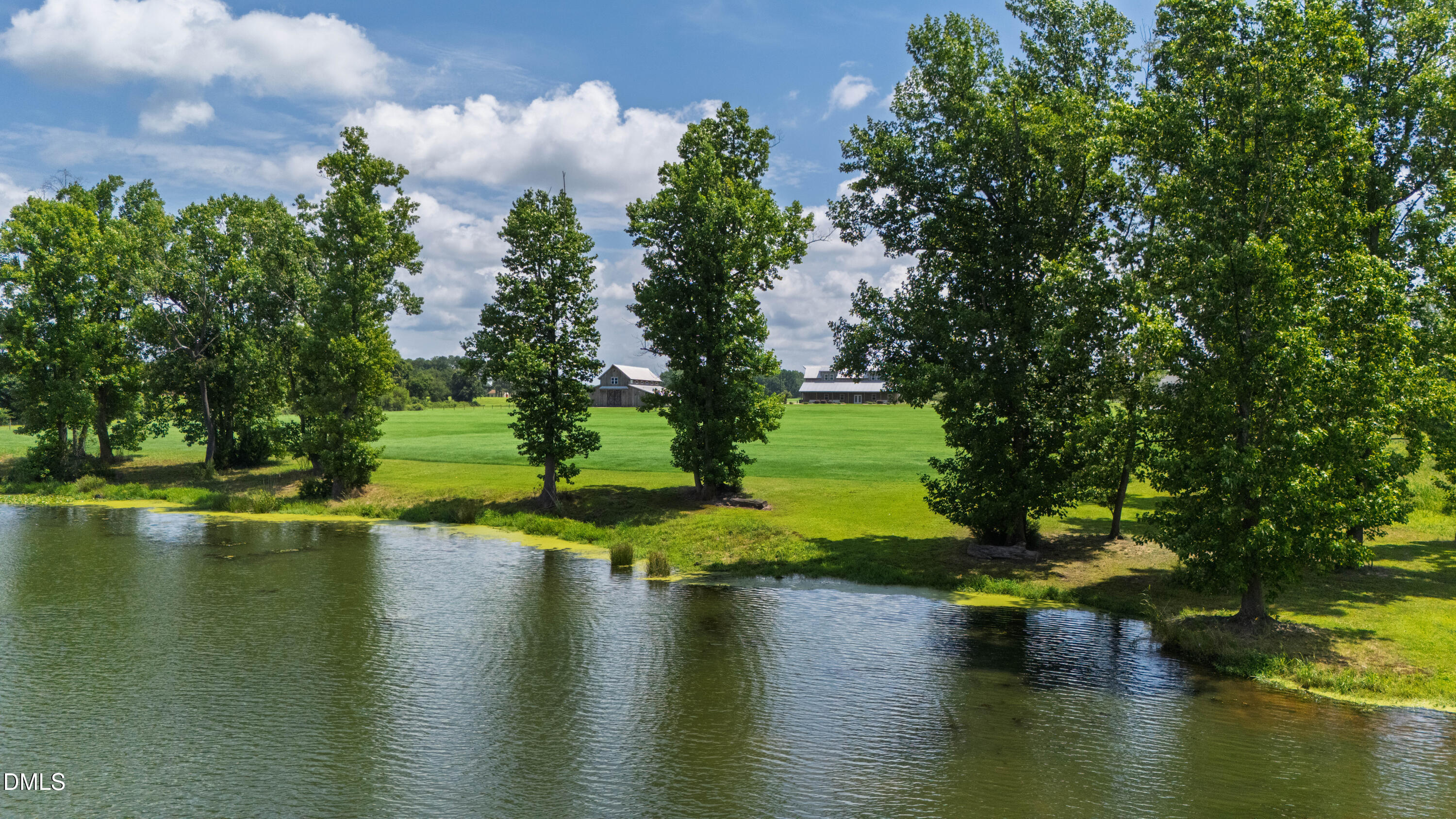 A pond borders a broad green lawn and mature trees, with farm buildings in the distance.