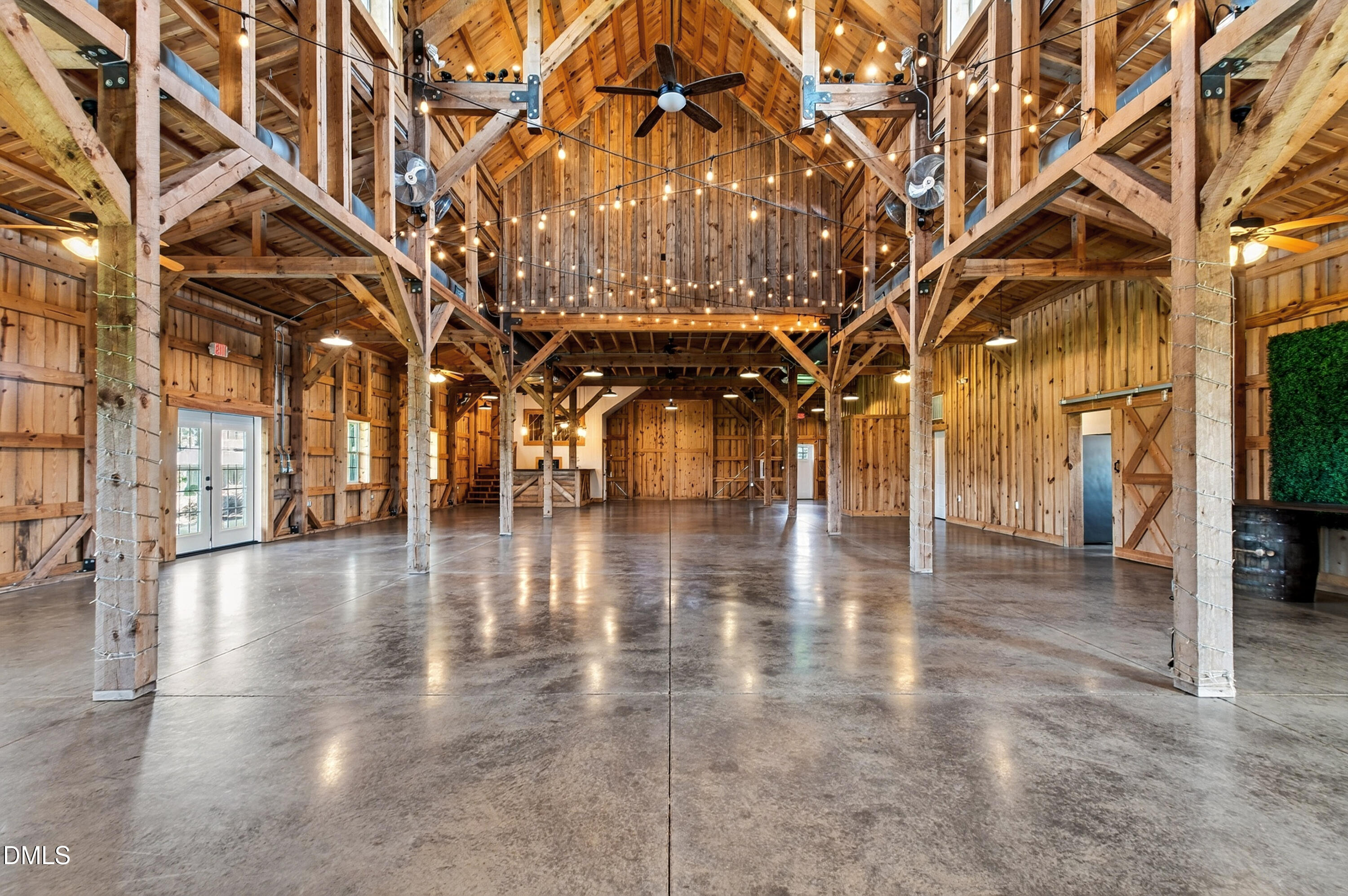 Open timber-frame barn interior with a vaulted ceiling, string lights, mezzanine, and polished concrete floor.