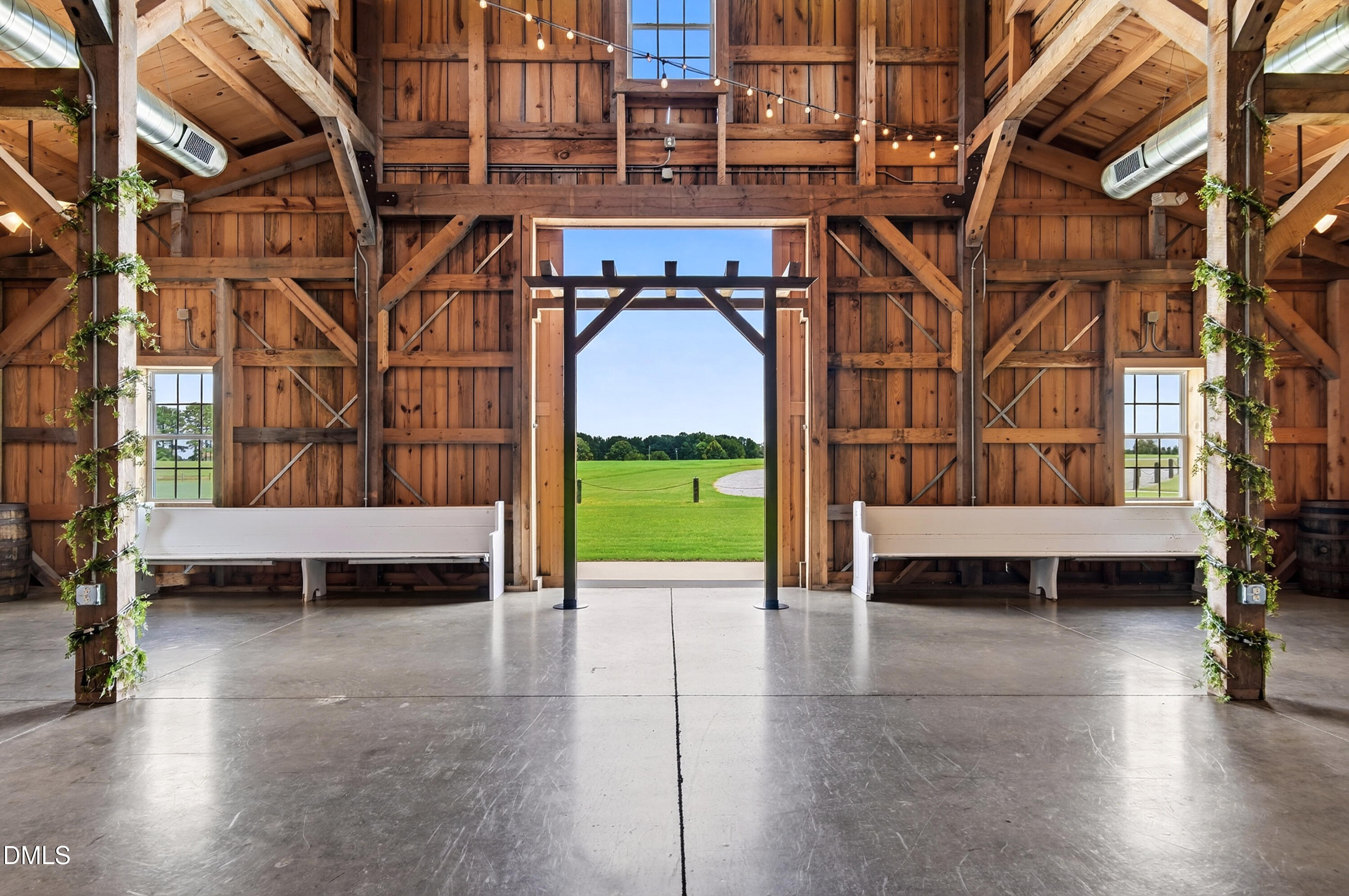 Timber-frame barn interior with white benches and a large open doorway overlooking a green lawn.