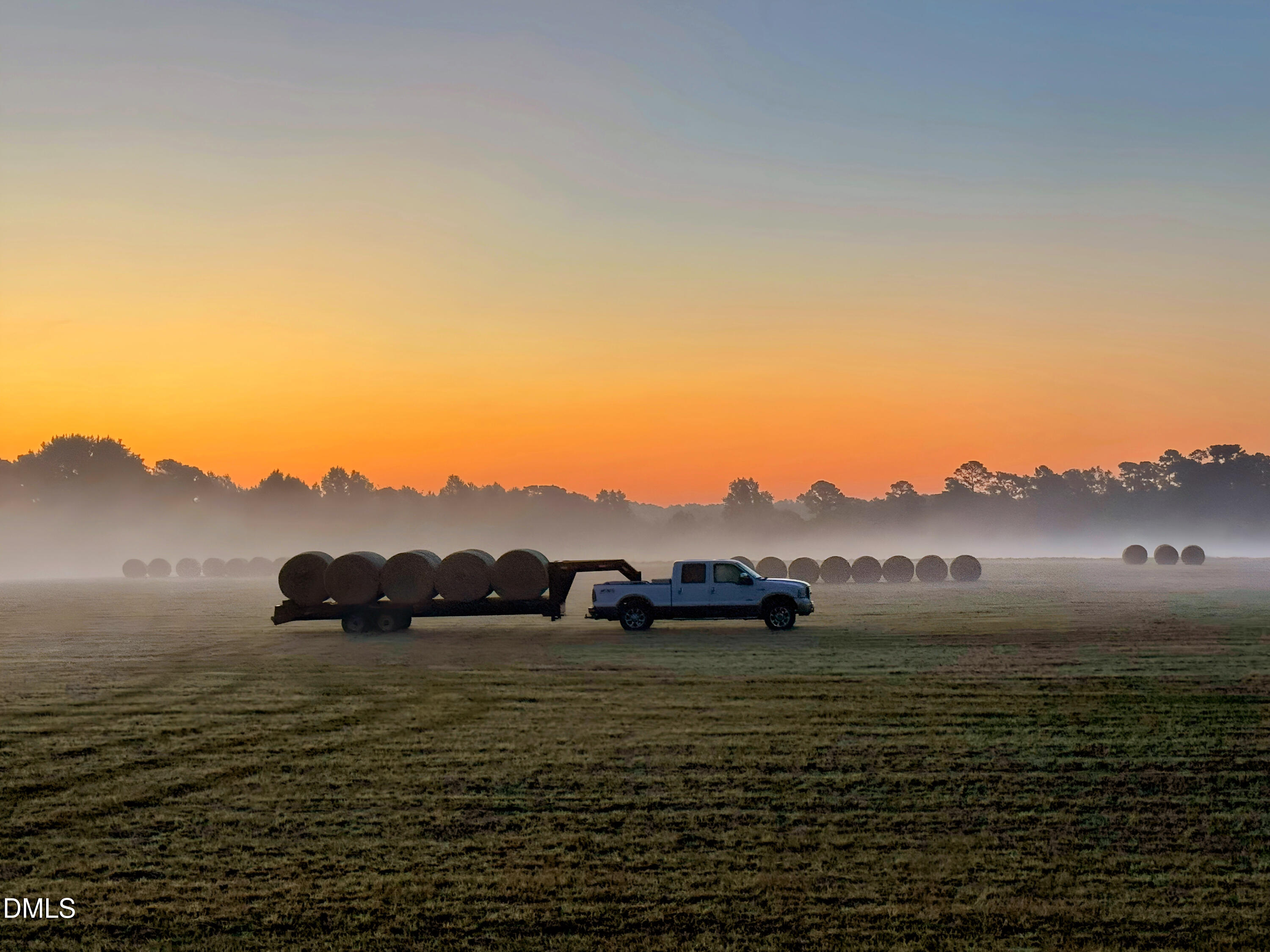 Open field with round hay bales and a truck at sunset.