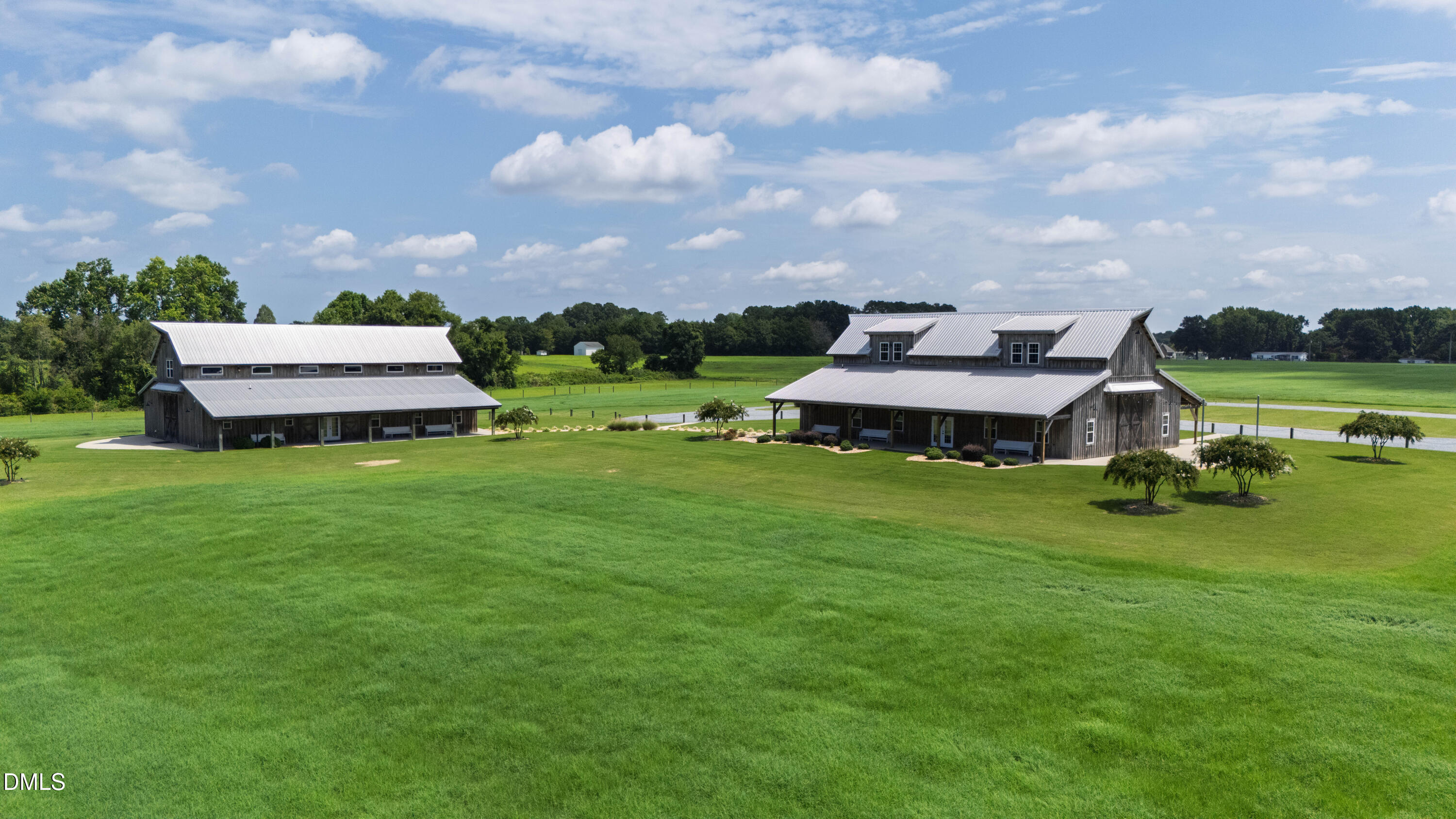 Two barn-style buildings with metal roofs stand across a broad green lawn beside open fields.