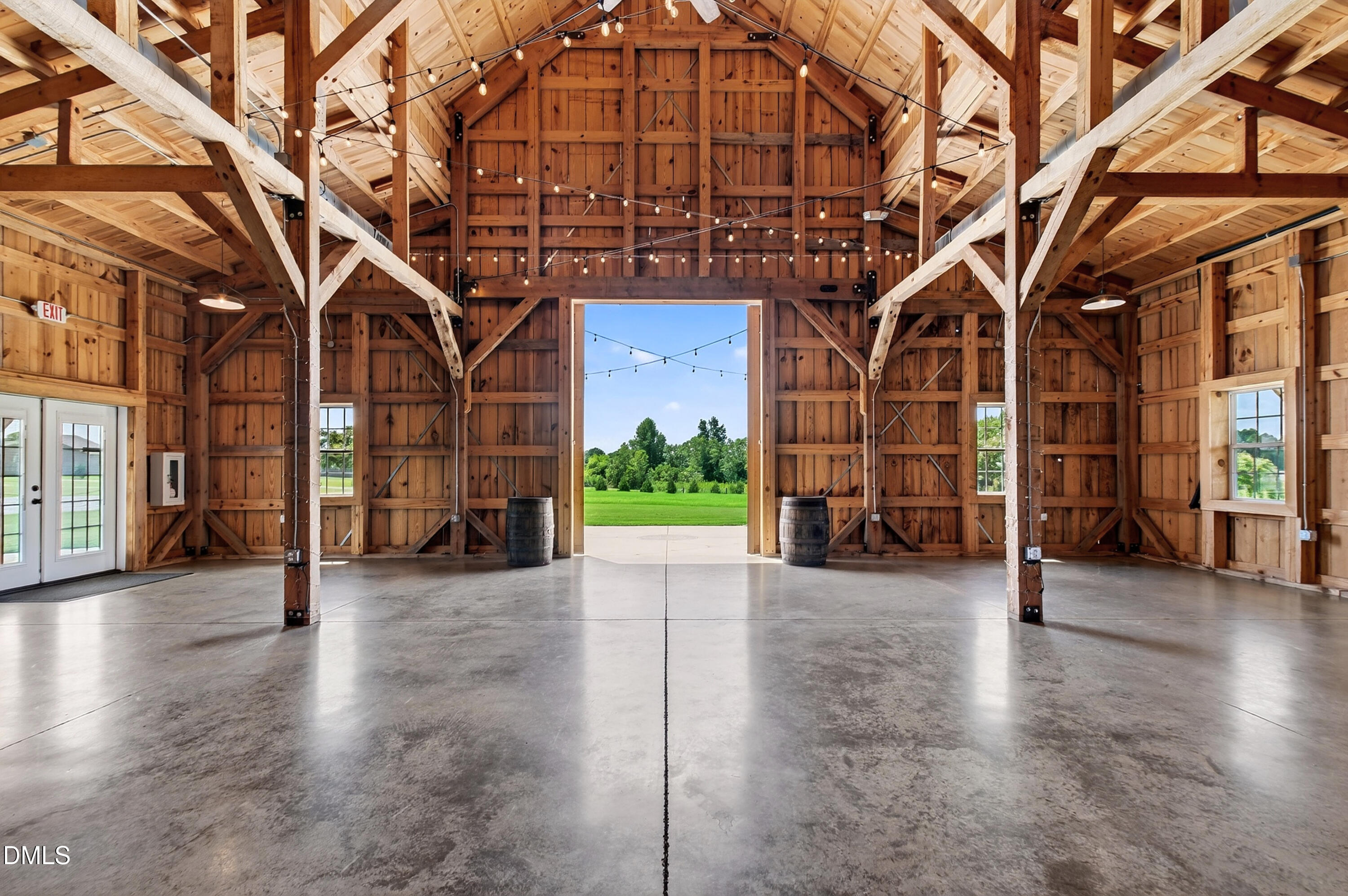 Timber-frame barn interior with vaulted beams, string lights, concrete floor, and open doorway onto a lawn.