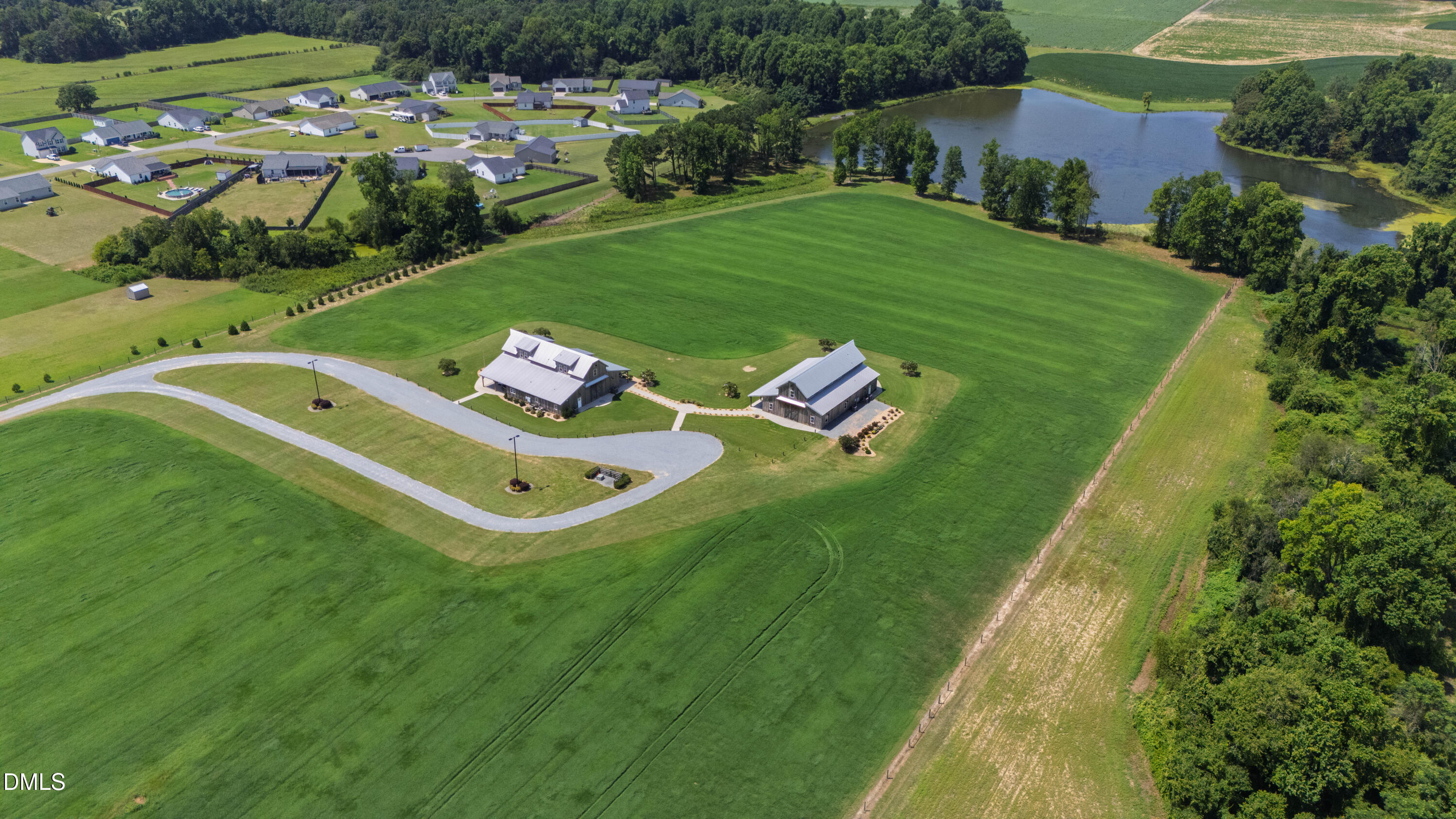 Aerial view of two buildings and a curving driveway amid green fields beside a pond.