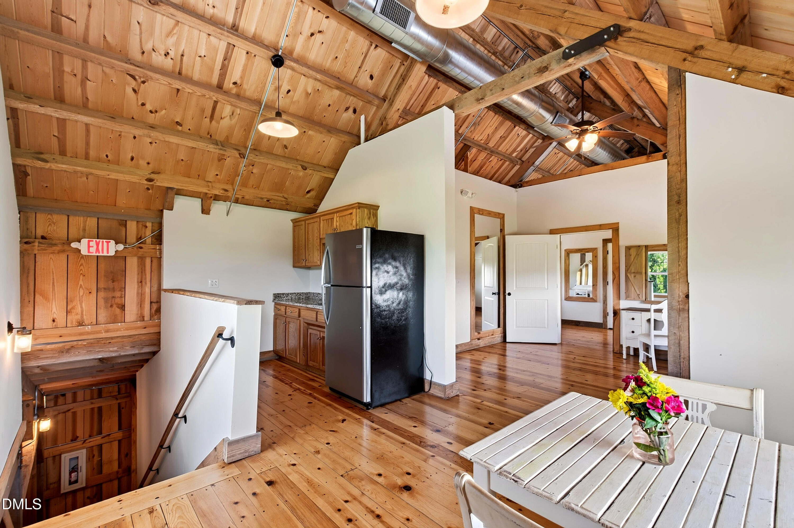 Rustic open kitchen with exposed timber ceiling, wooden cabinets, and a white dining table beside the stairs.