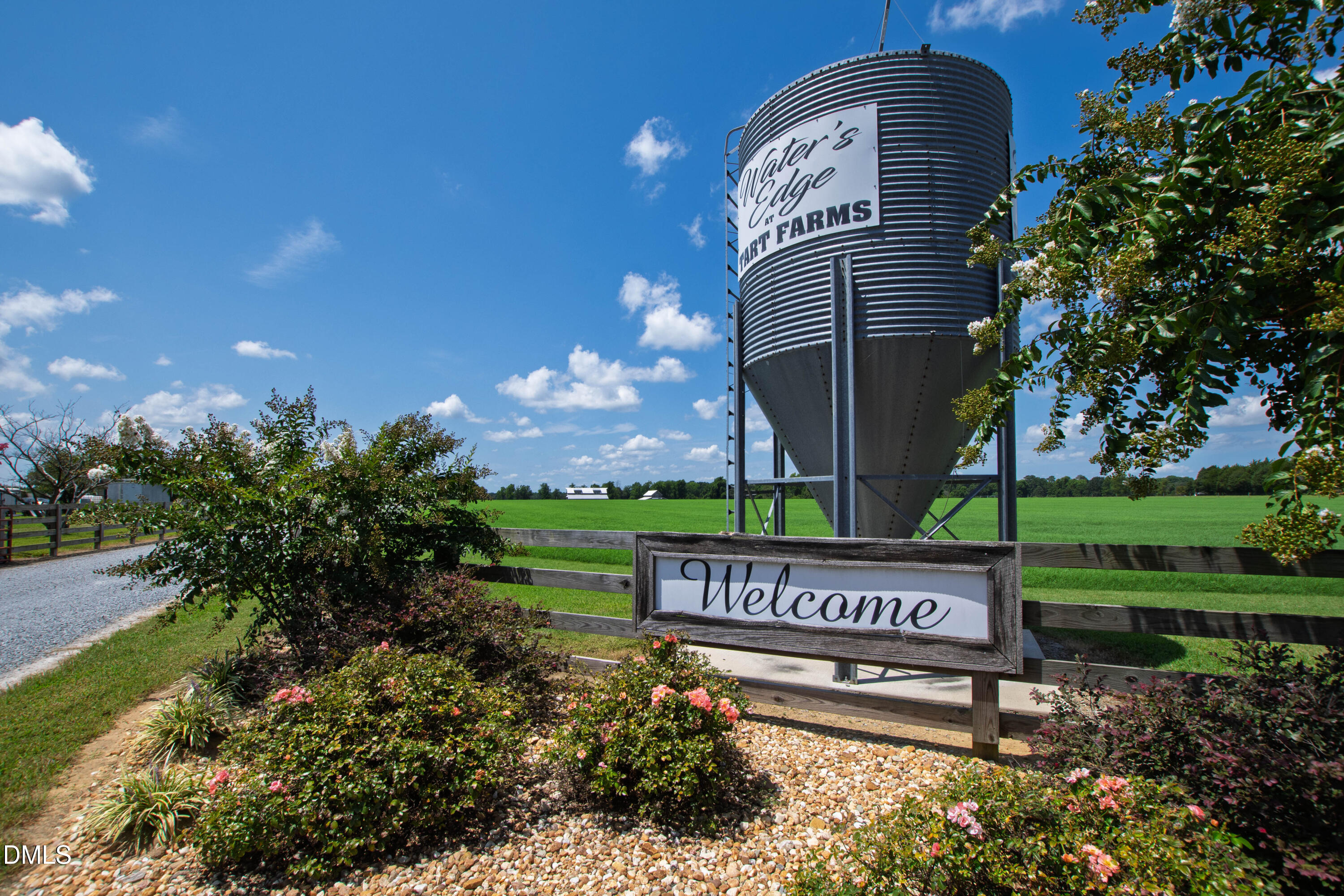 Farm entrance with a Welcome sign, flower beds, green fields, and a silo marked Water’s Edge at Hart Farms.