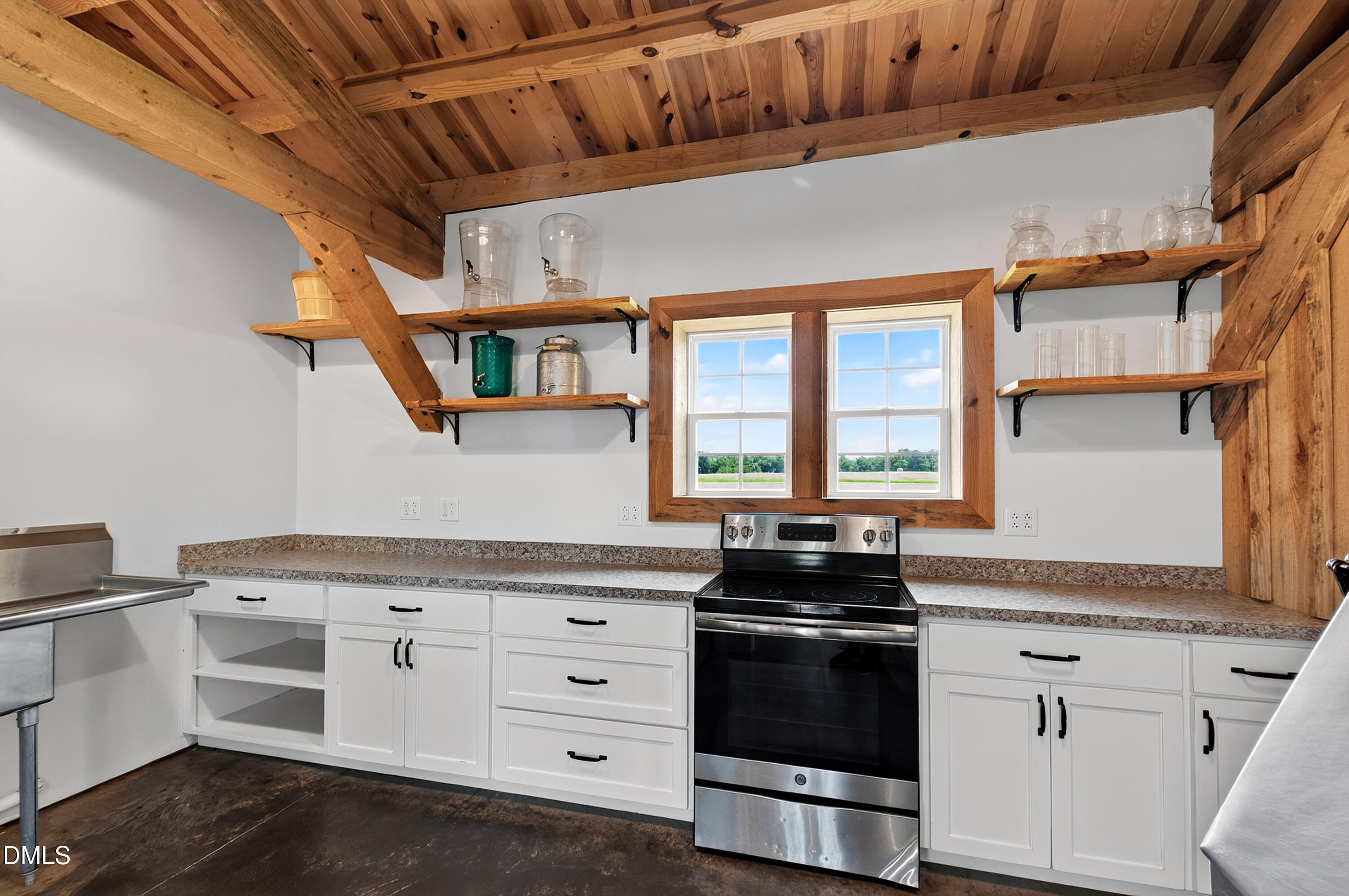 Kitchen with exposed timber ceiling, open wood shelves, white cabinets, and a range beneath a double window.