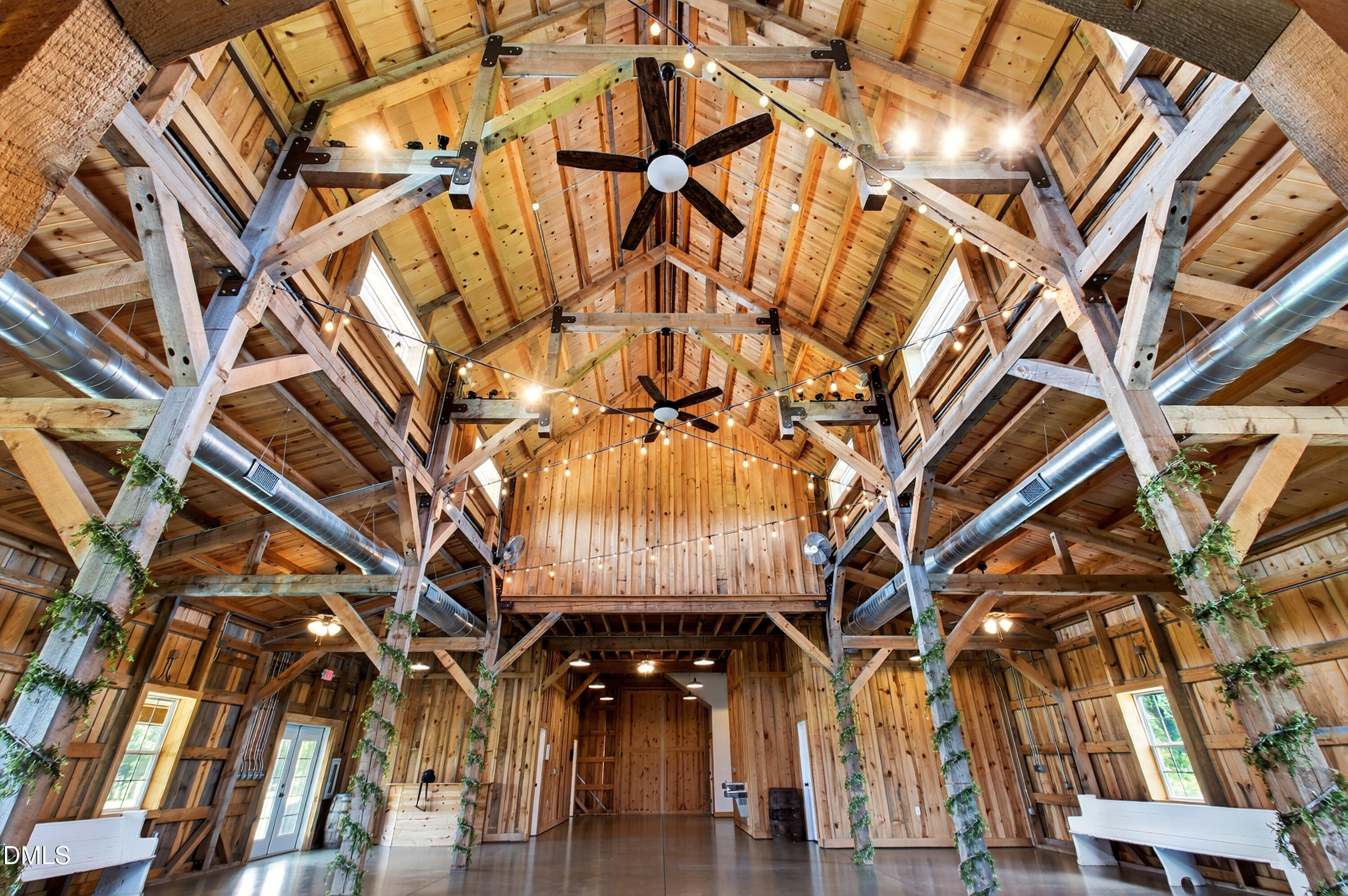 Timber-frame barn interior with vaulted wooden ceiling, exposed beams, ceiling fans, and string lights.