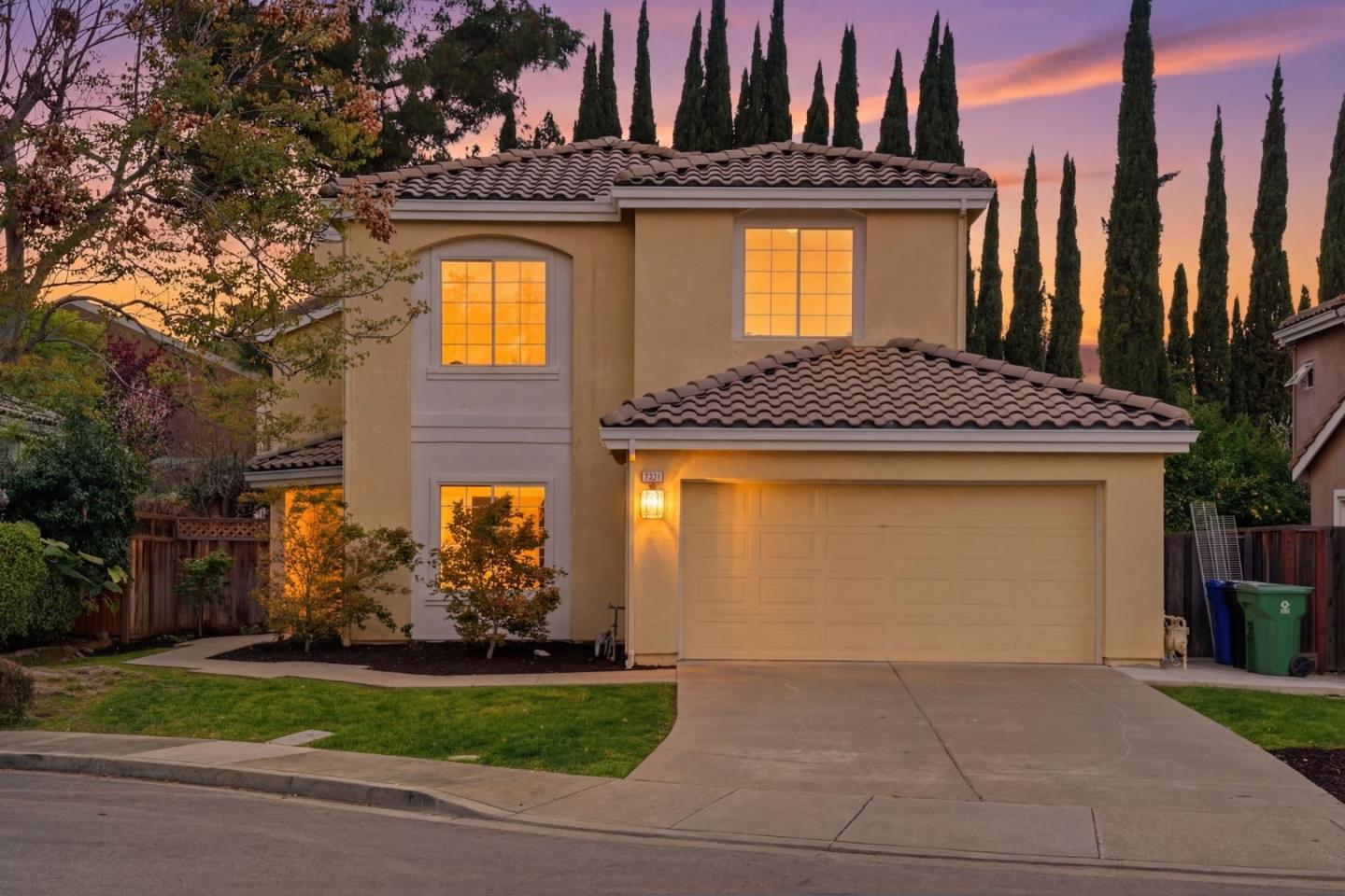 Two-story stucco home with a tiled roof and glowing windows beneath a pink-orange dusk sky.