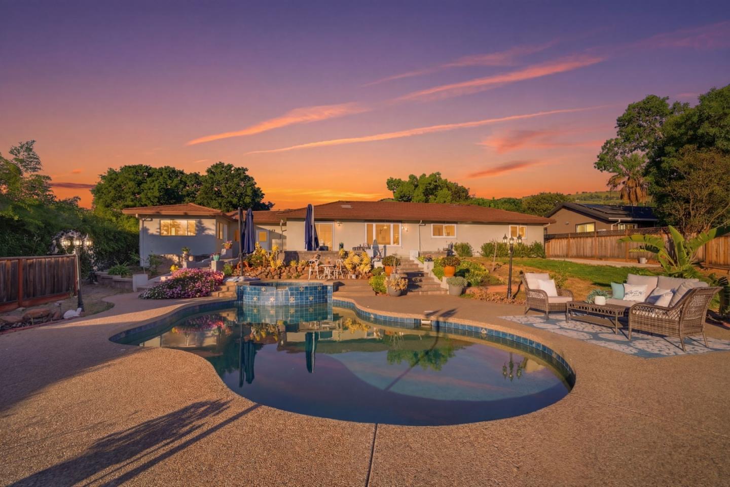Single-story home behind a curved backyard pool, patio seating, and garden beds at sunset.