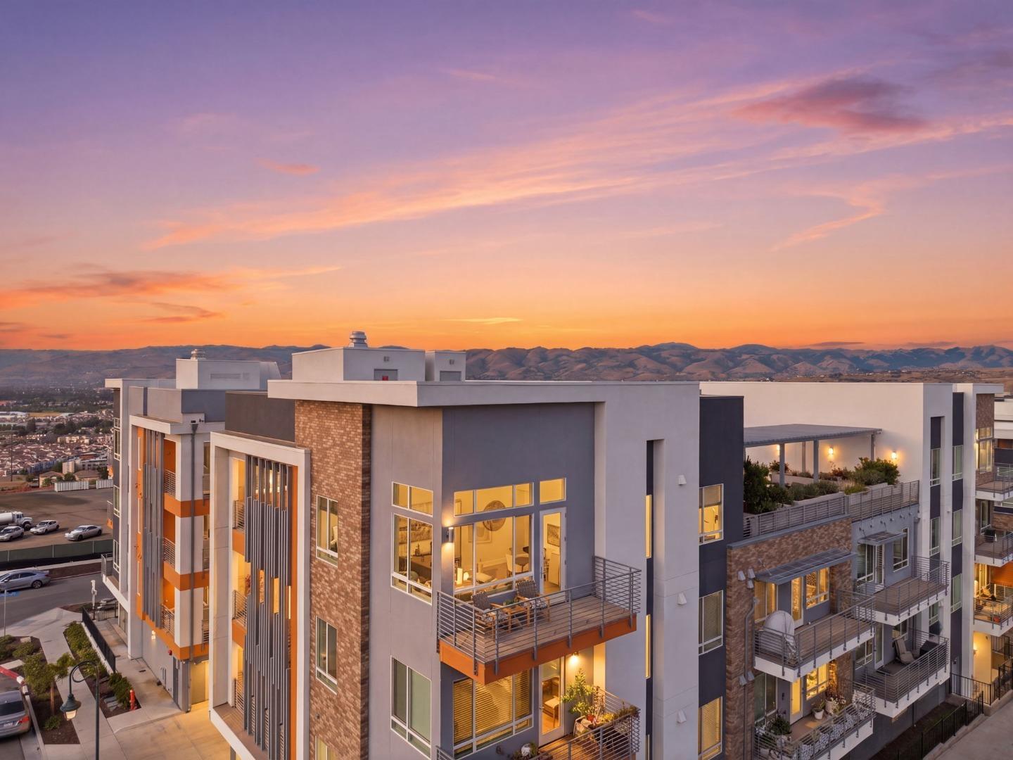 Modern residential building with balconies and glowing windows beneath a sunset sky, with hills in the distance.