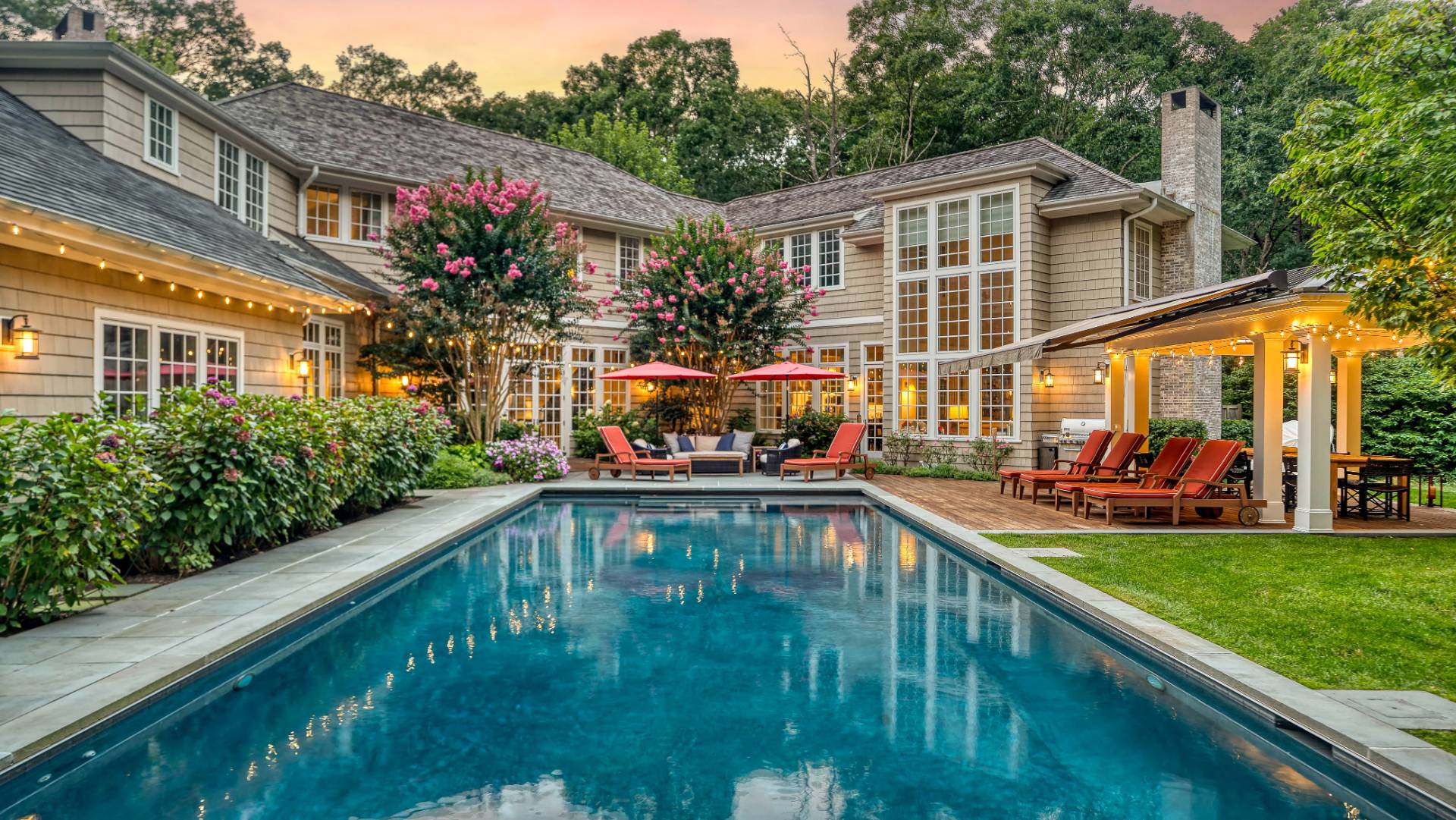 Shingle-style home behind a swimming pool, with flowering trees and a covered patio at dusk.
