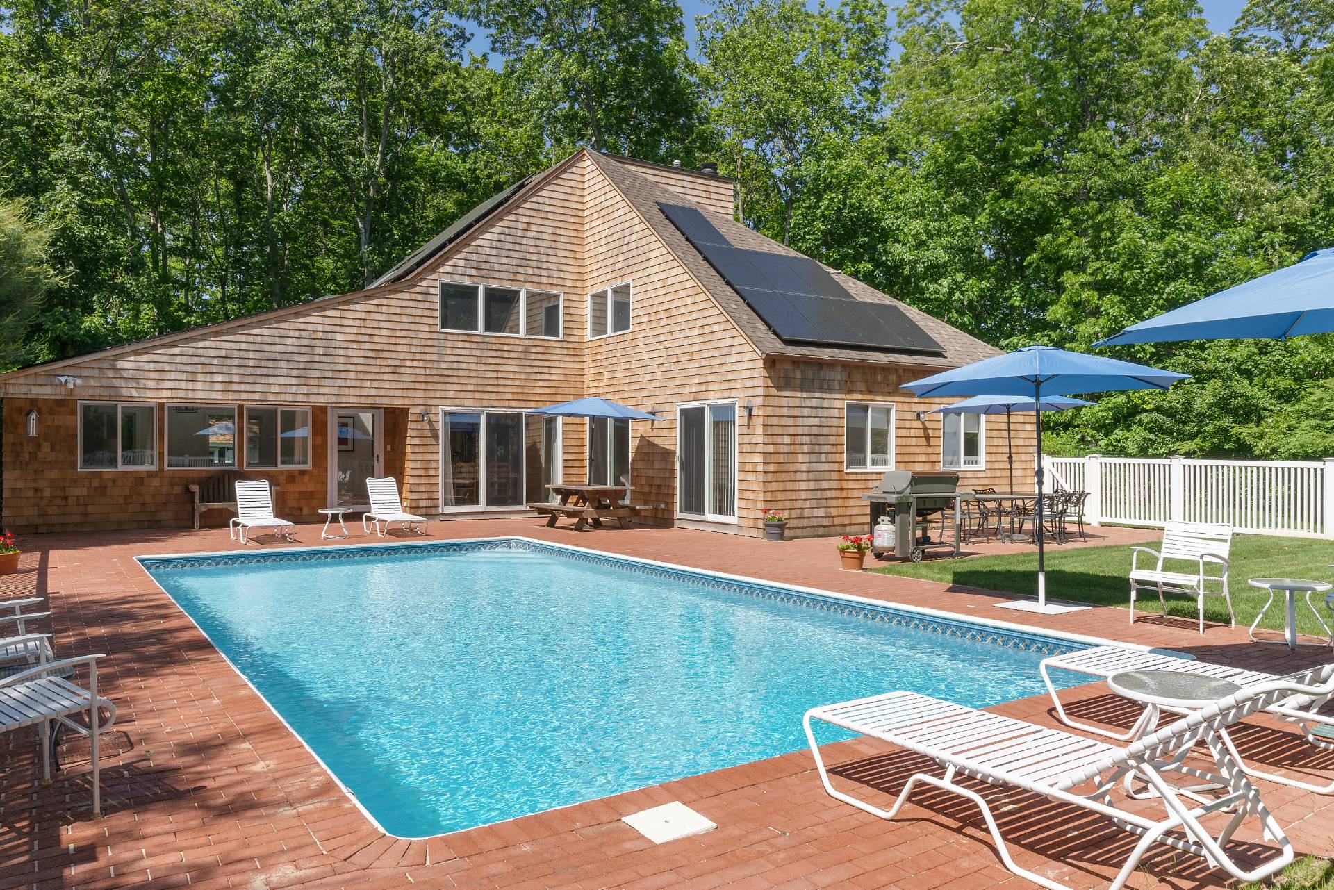 Cedar-shingle house with rooftop solar panels behind a swimming pool and furnished brick patio.