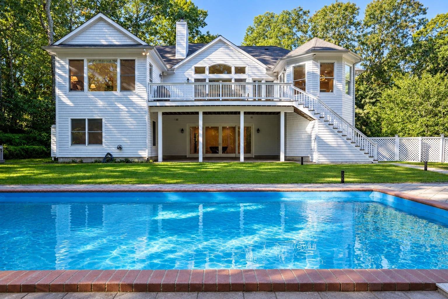 White two-story house with a raised deck and exterior staircase behind a large swimming pool.