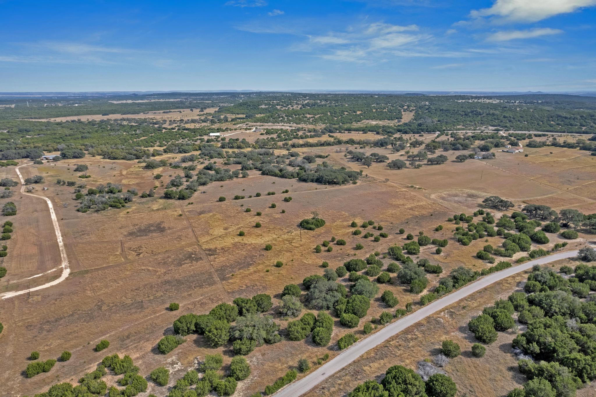 Aerial view of dry rural acreage with scattered trees and a paved road along the foreground.