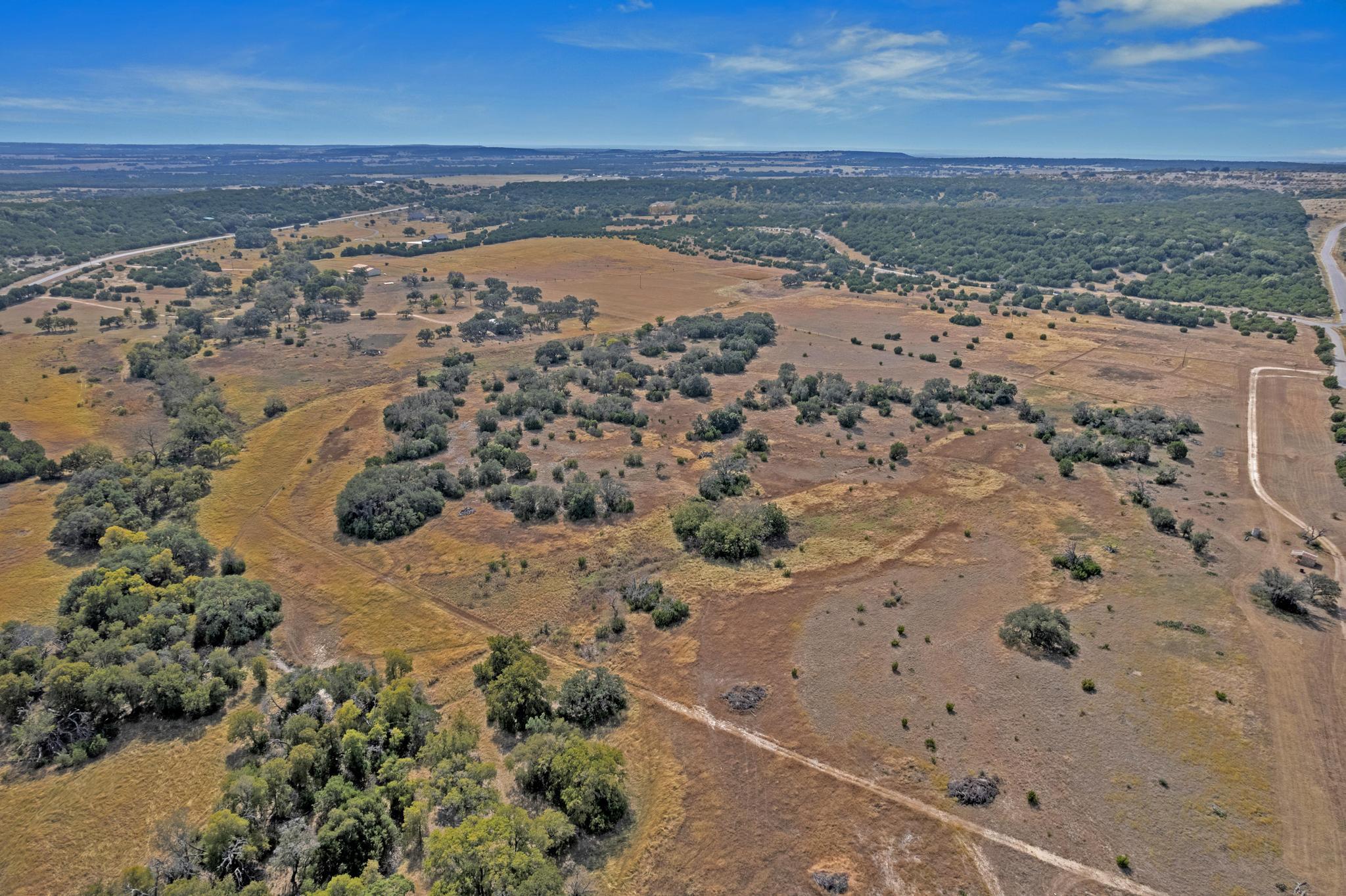 Aerial view of dry ranchland dotted with trees and crossed by dirt tracks, with a road at the right edge.