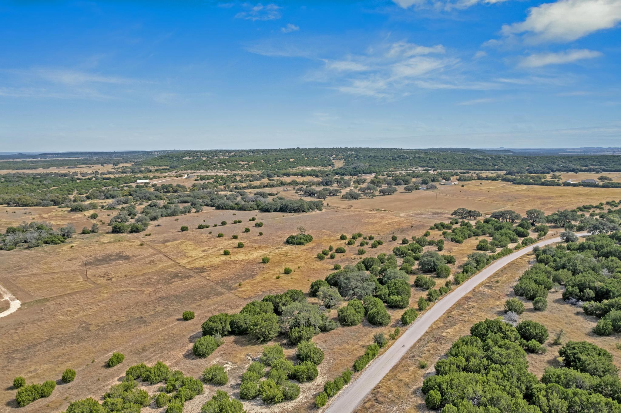 Aerial view of dry Texas ranchland with scattered trees and a paved road winding through it.