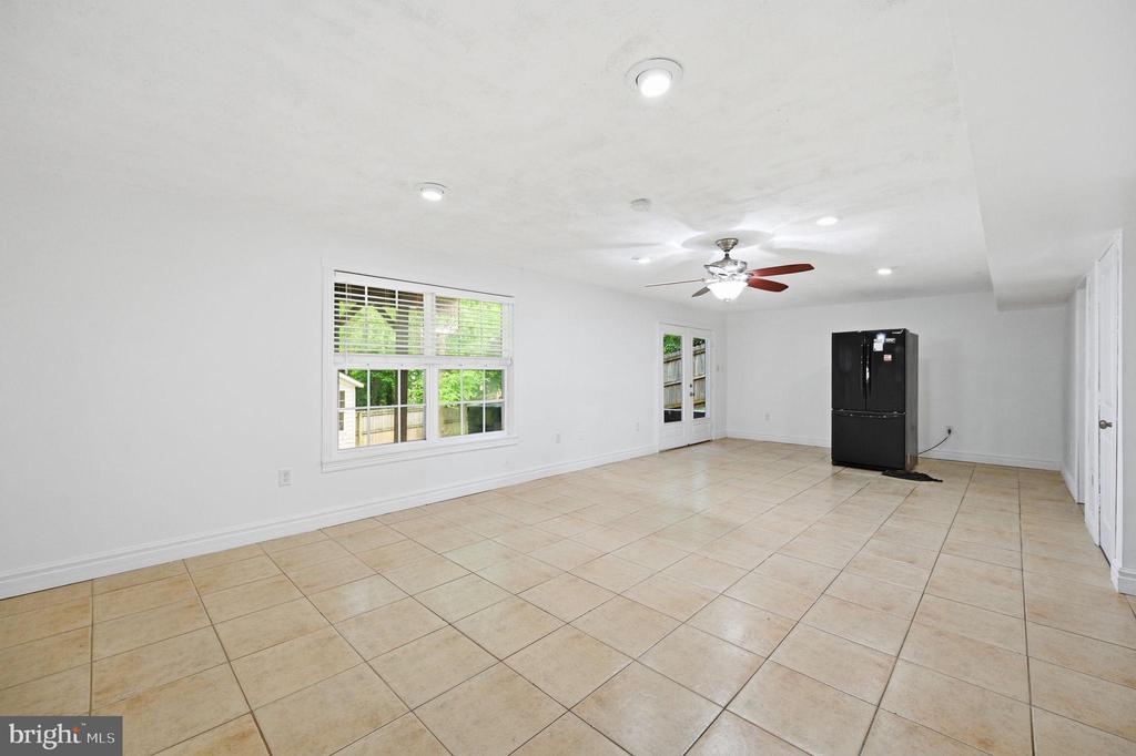 Empty tiled room with a ceiling fan, large windows, glass doors, and a black refrigerator.