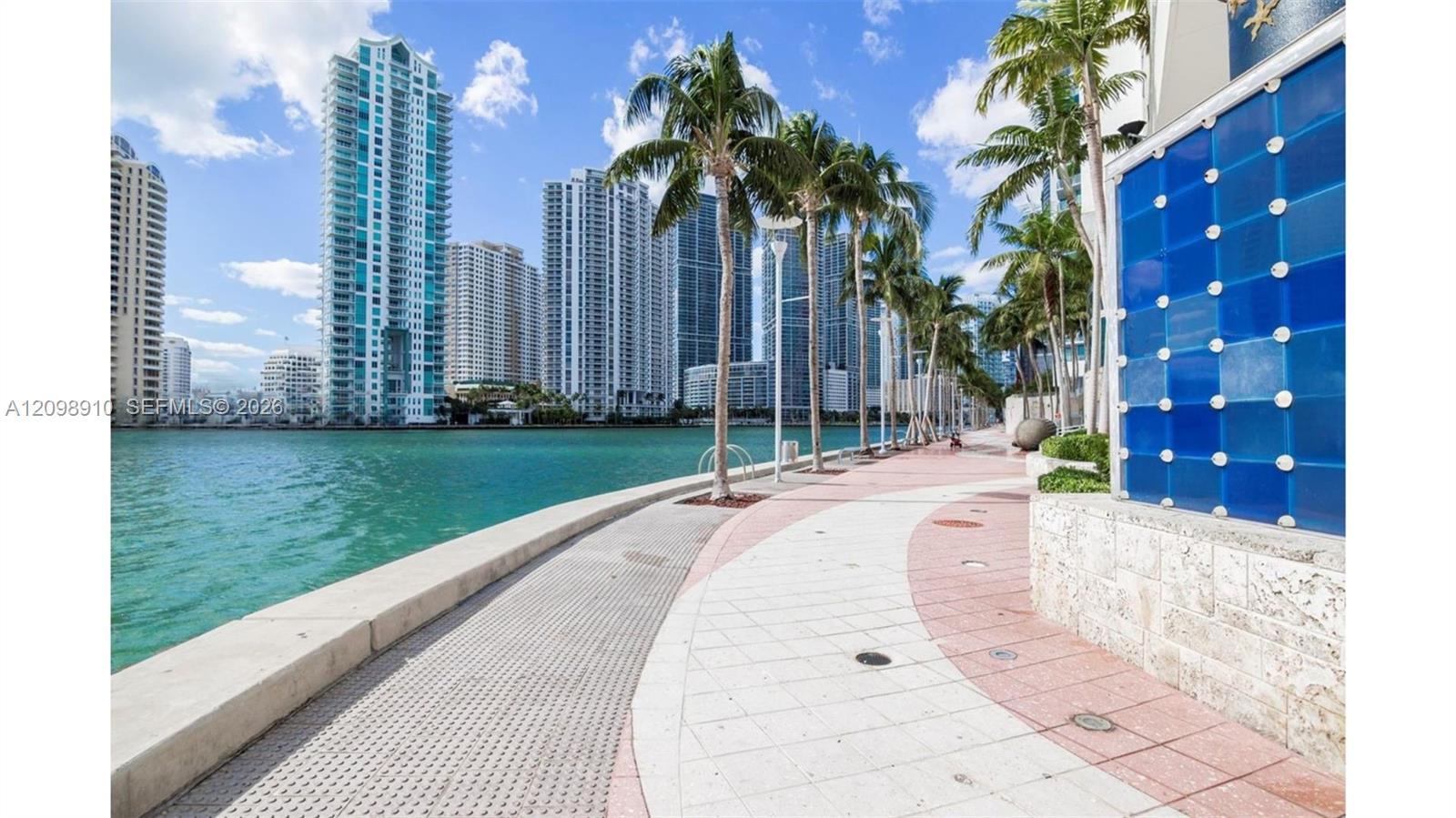 Waterfront promenade with palm trees, turquoise water, and Miami high-rises across the bay.