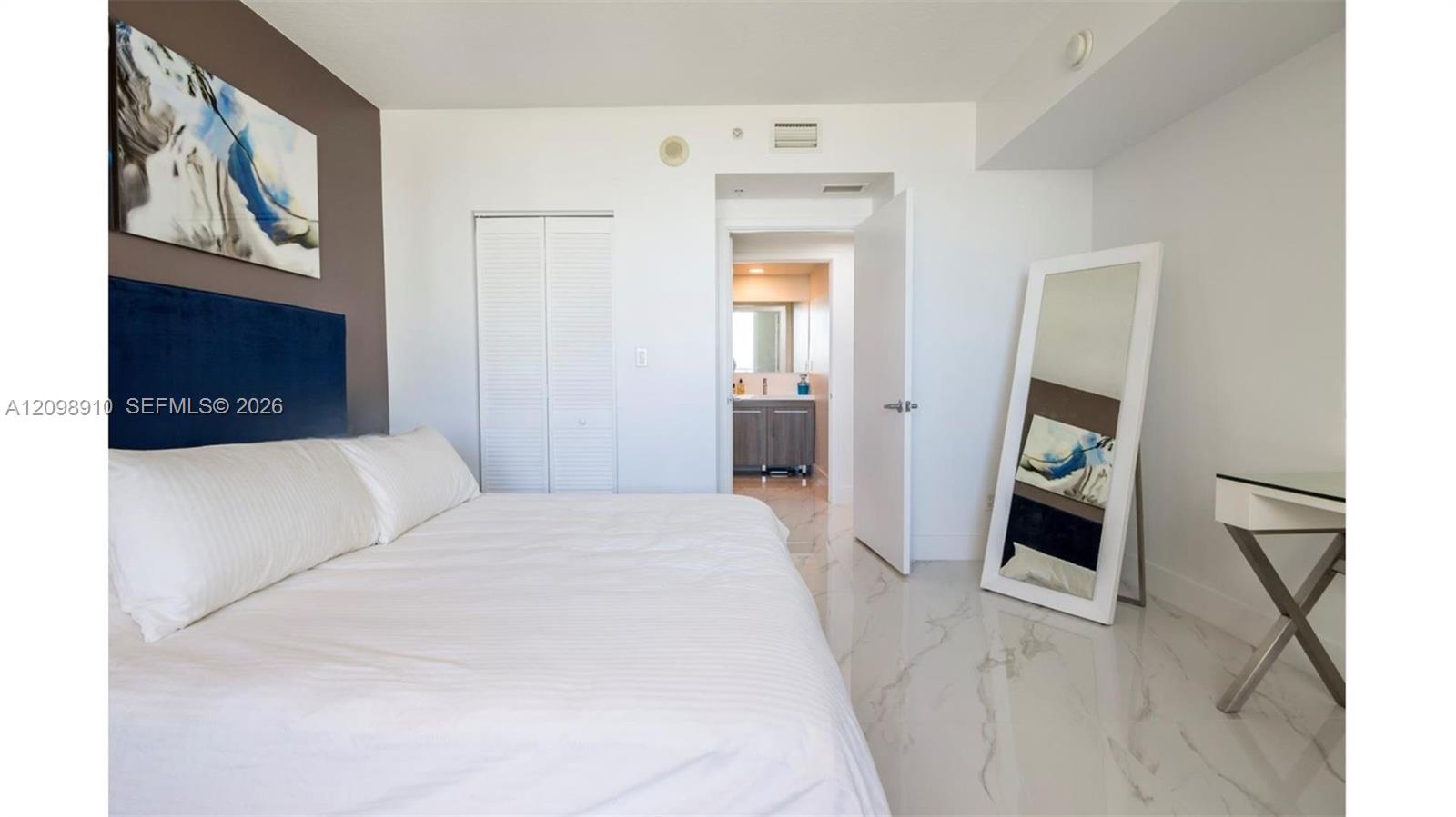 Furnished bedroom with a white bed, navy headboard, marble-look floors, and an open bathroom doorway.