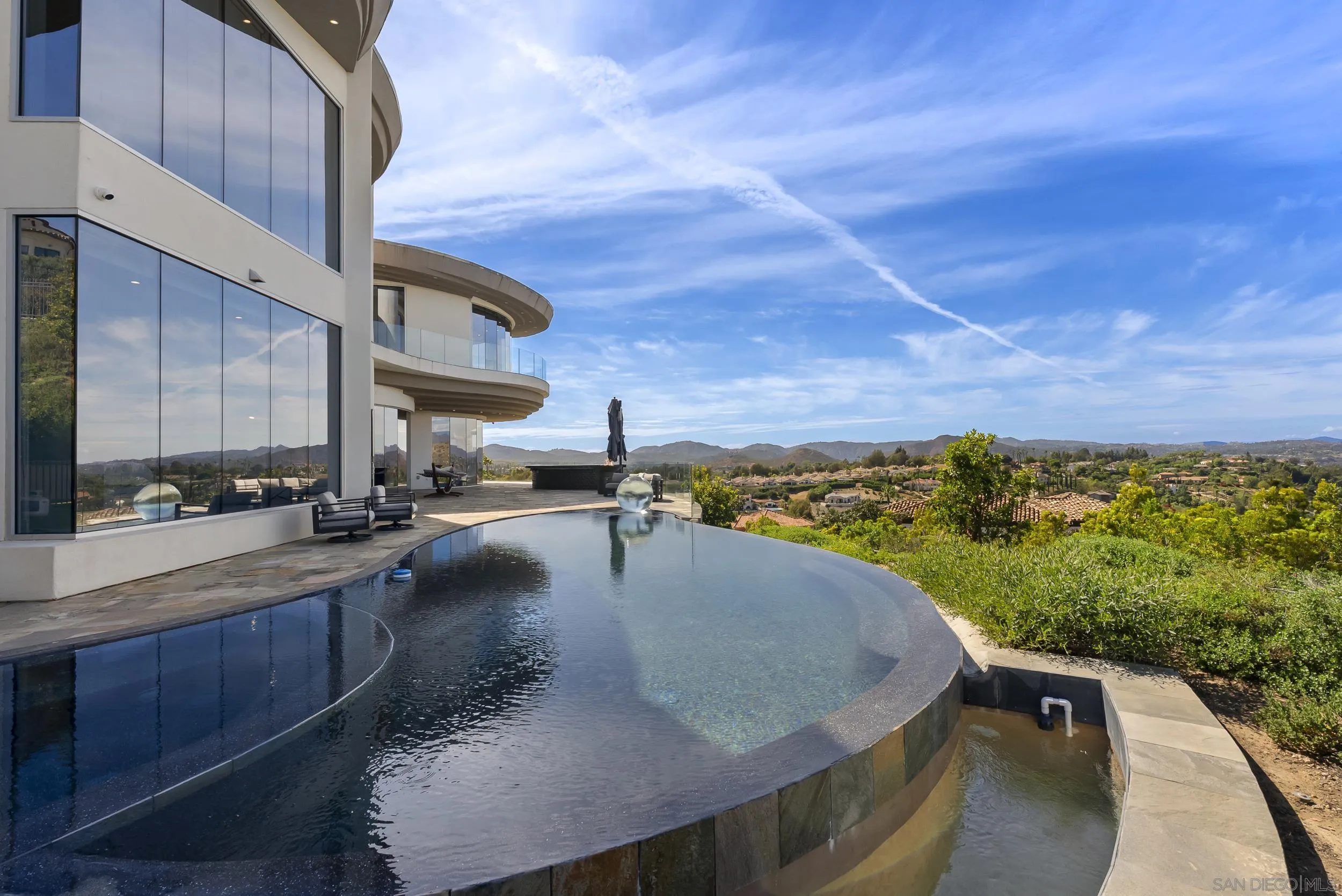 Modern glass home beside a curved infinity pool overlooking a hillside neighborhood and distant mountains.