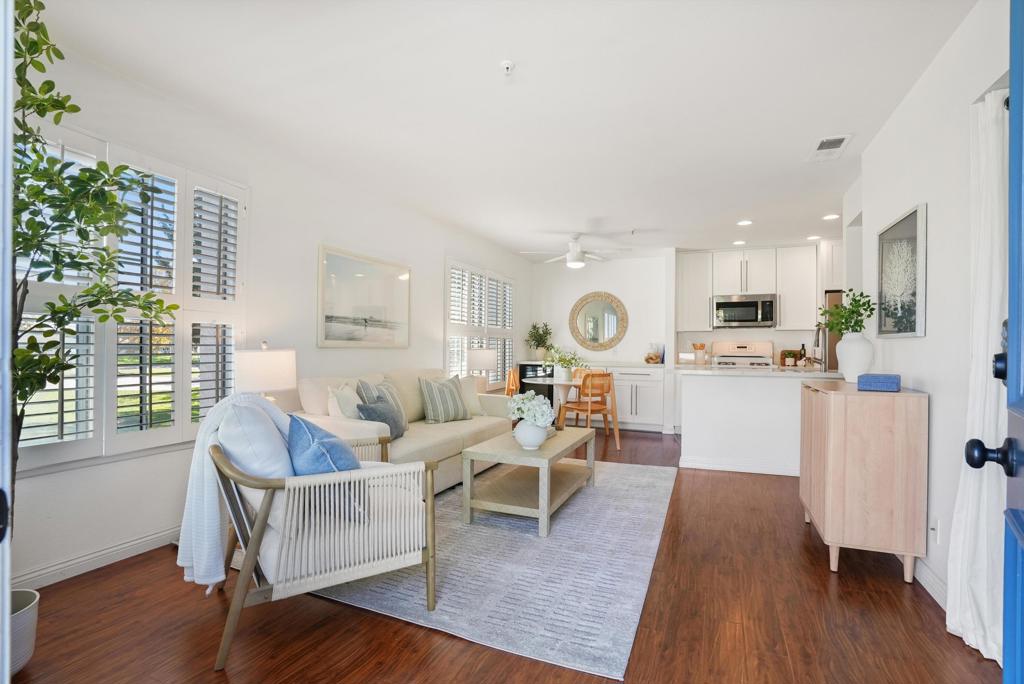 Furnished living room with plantation shutters, hardwood floors, and an open white kitchen.