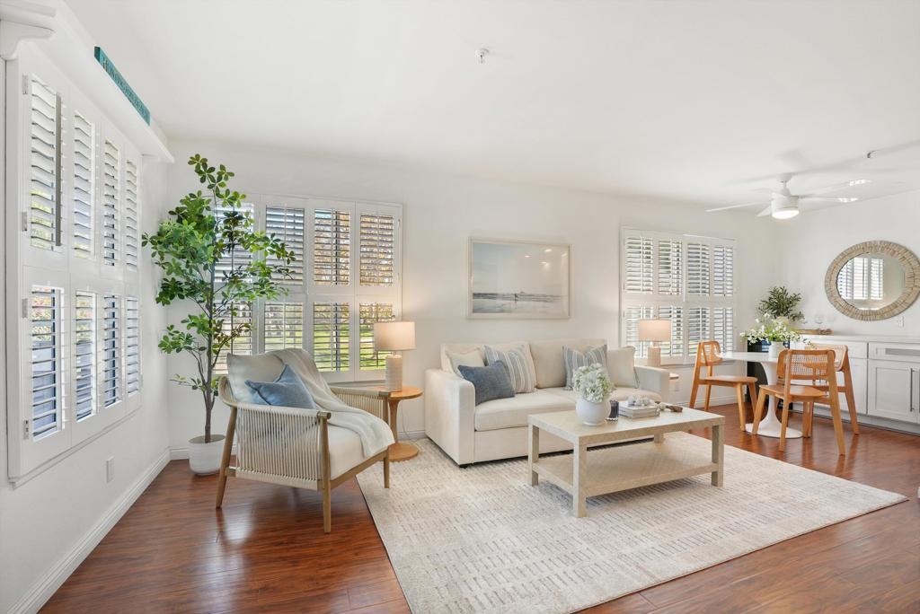 Staged living room with plantation shutters, white seating, hardwood floors, and an adjoining dining area.