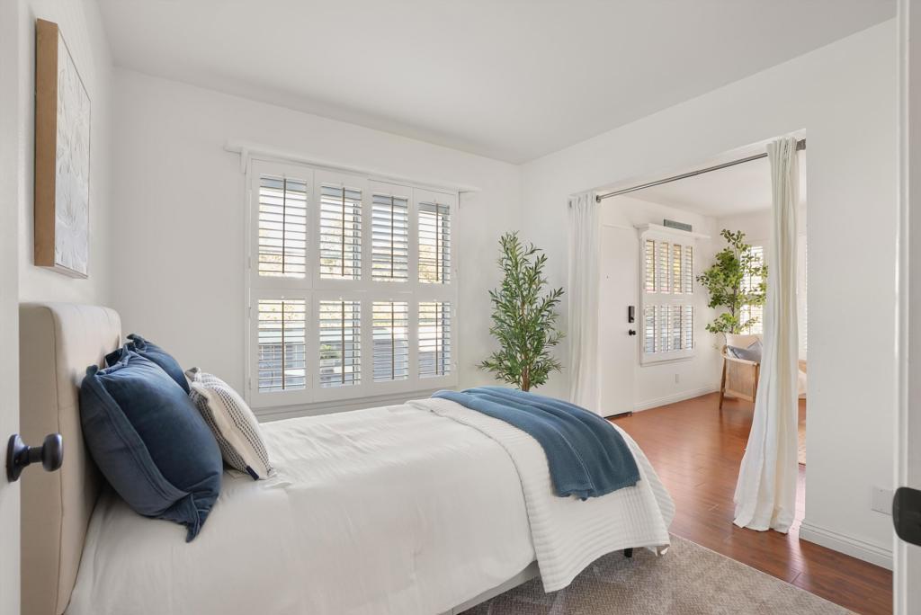 Staged bedroom with a white bed, navy accents, plantation shutters, and an adjoining sitting area.
