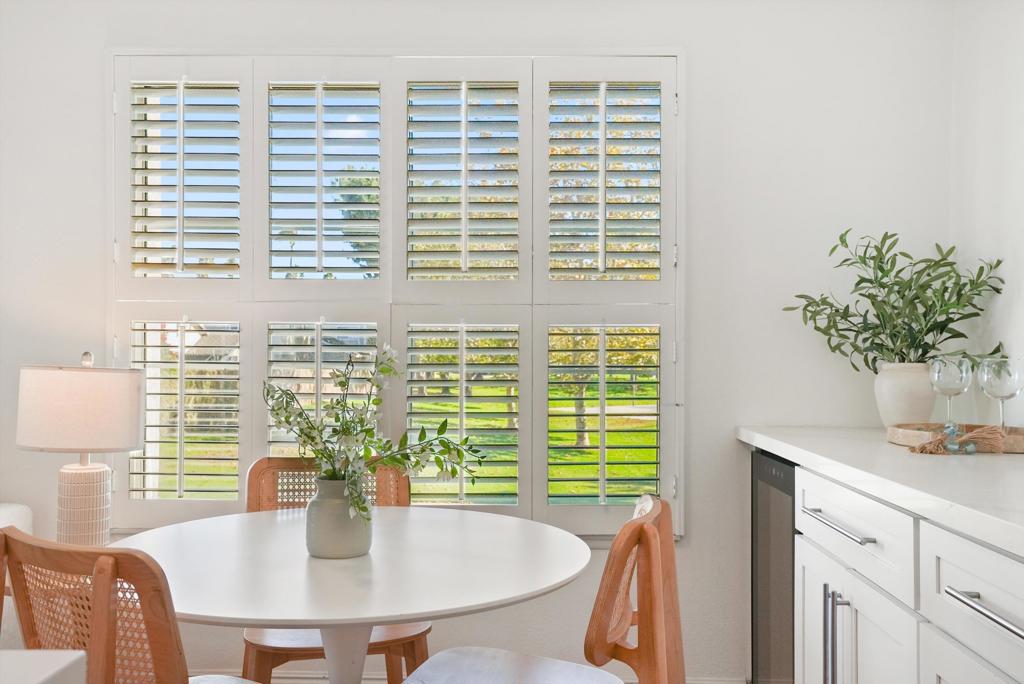 Dining area with a round white table, woven chairs, and plantation shutters overlooking a green lawn.