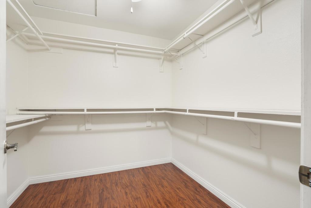 Empty walk-in closet with wraparound white shelves and hanging rods over wood-look flooring.