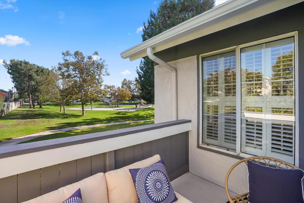 Apartment balcony overlooking a green park, with cushioned seating and plantation-shutter windows.