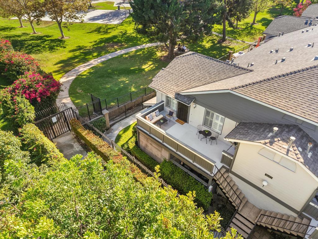 Aerial view of a home’s furnished patio beside a landscaped park with a curving path and flowering shrubs.