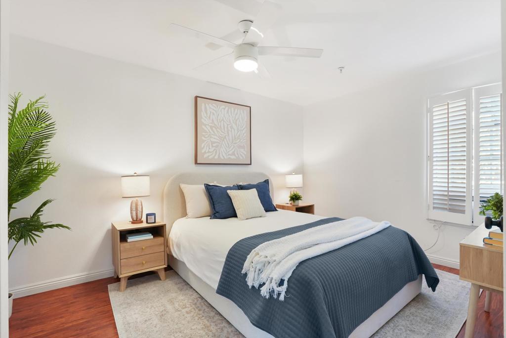 Staged bedroom with a blue quilt, white throw, plantation shutters, and a ceiling fan.