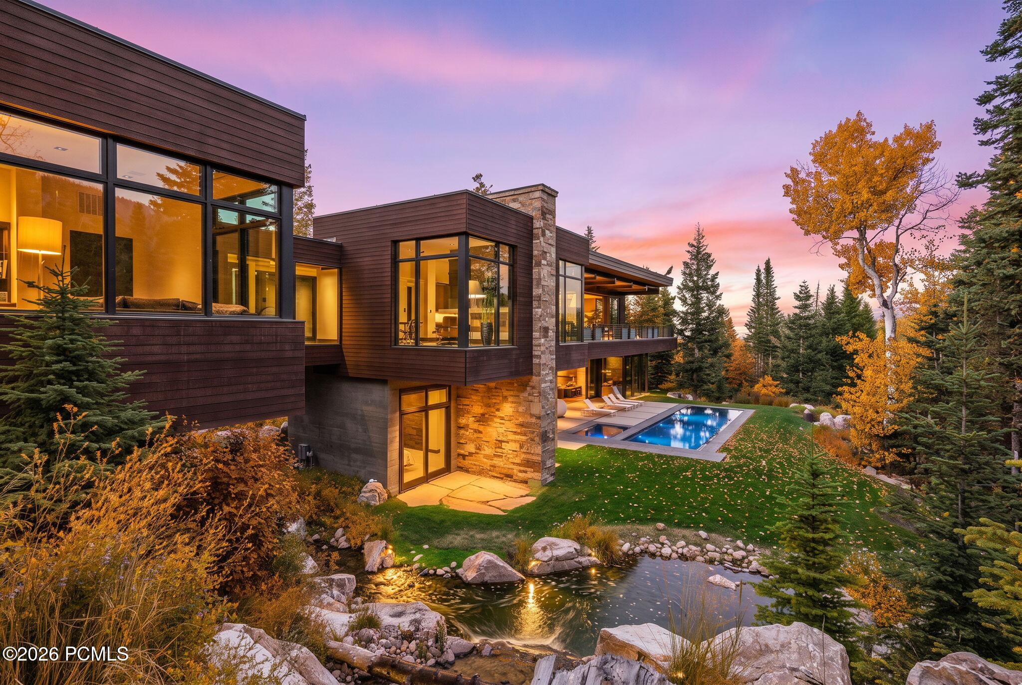 Modern home with glowing windows, a swimming pool, and landscaped pond at autumn dusk.