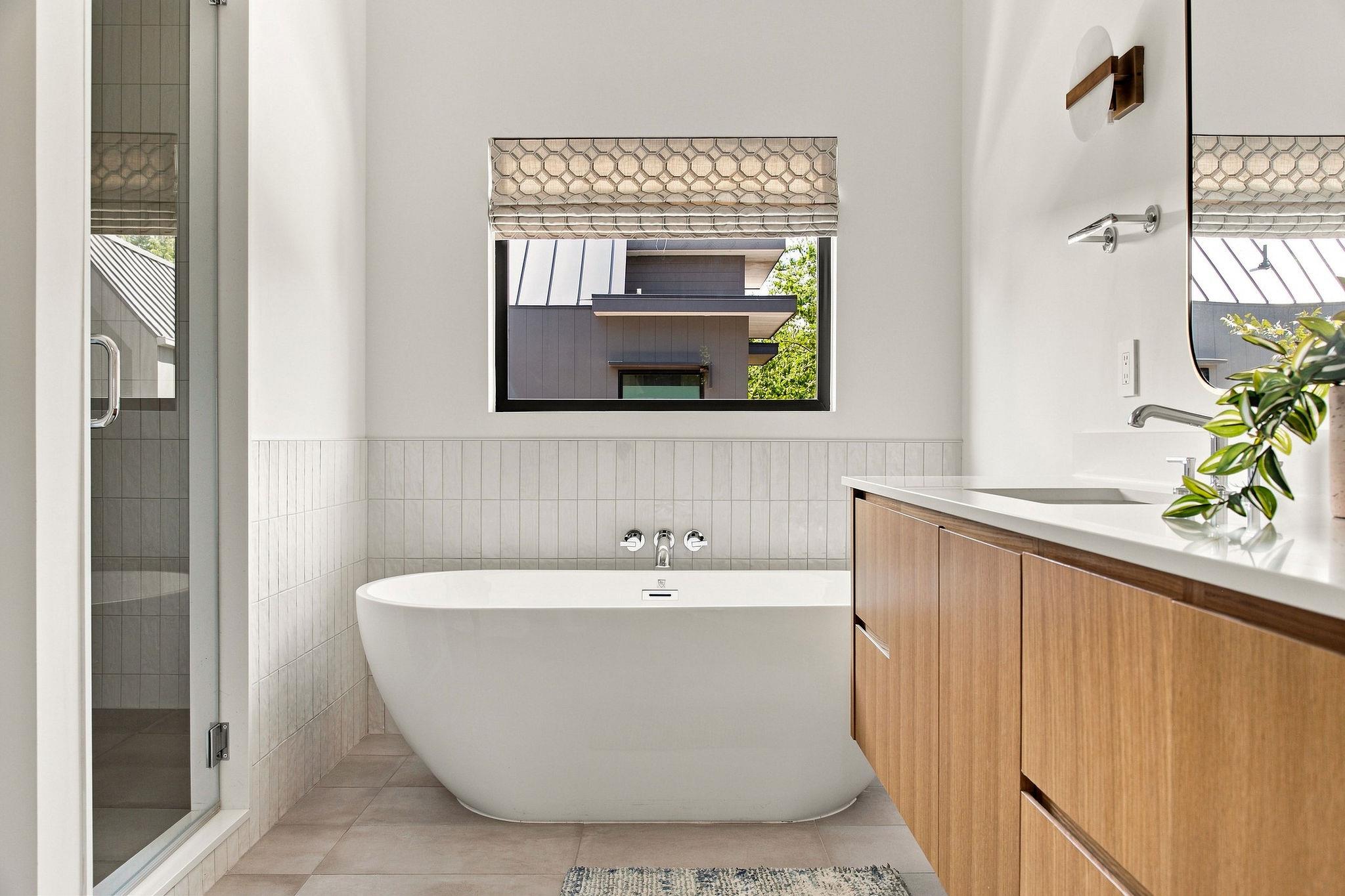 Modern bathroom with a freestanding soaking tub, wood vanity, and window facing a neighboring home.