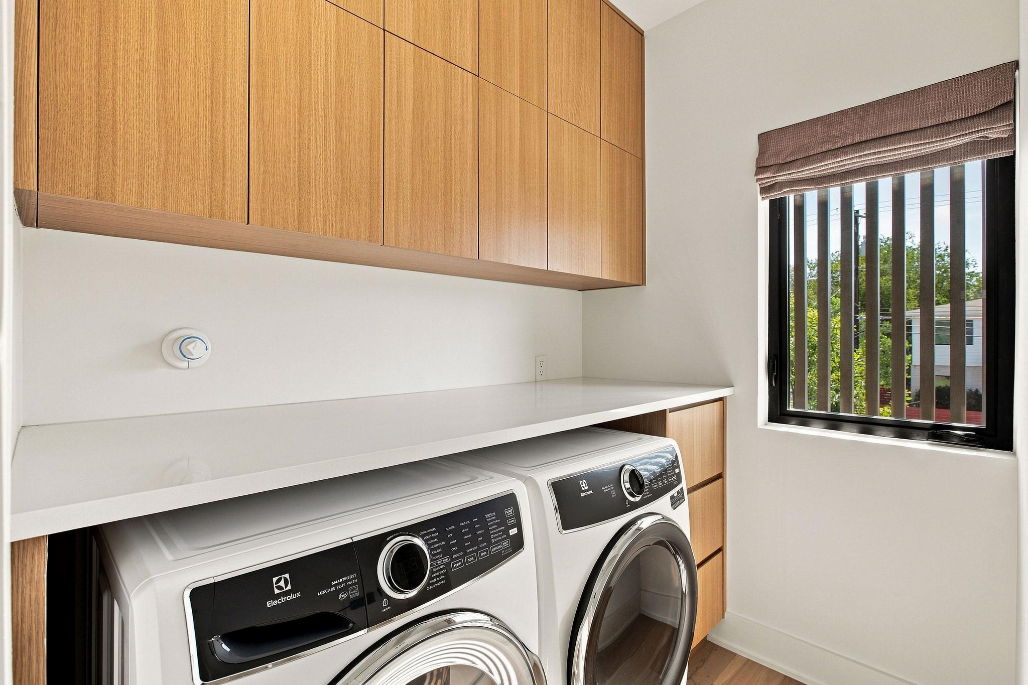 Laundry room with Electrolux washer and dryer, wood cabinets, and a window with a Roman shade.