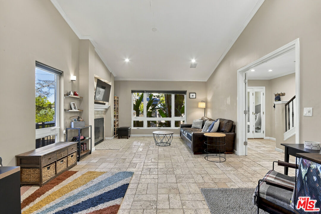 Living room with a leather sofa, tiled floors, fireplace, and large windows overlooking greenery.