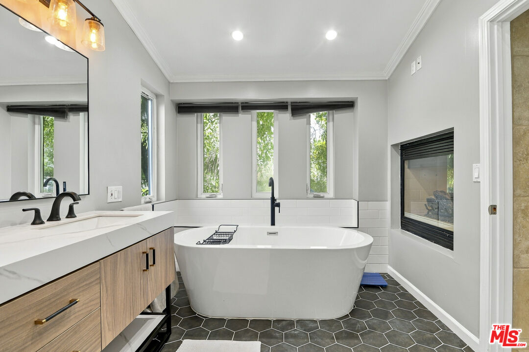 Modern bathroom with a freestanding tub beneath three windows and a built-in fireplace.