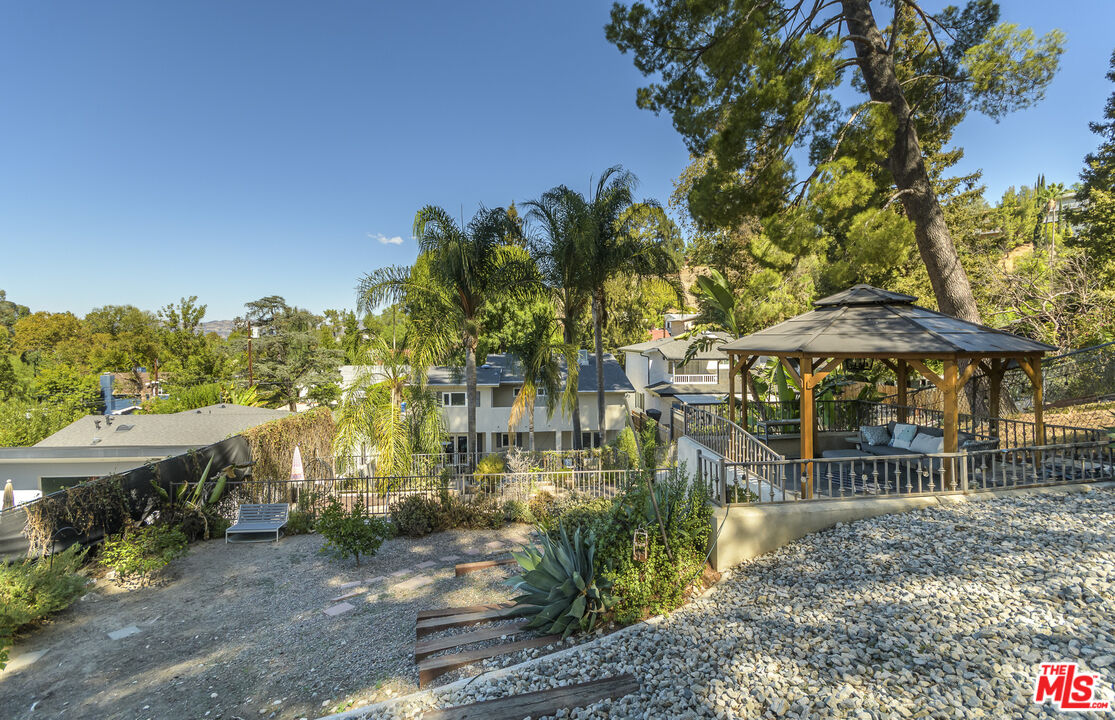 Backyard gazebo with seating beside a landscaped gravel yard and palm trees.