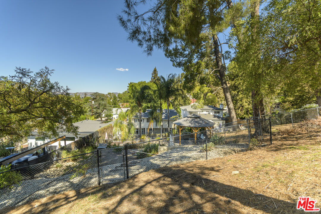 Home below a wooded hillside, viewed over a chain-link fence with a covered gazebo and distant hills.