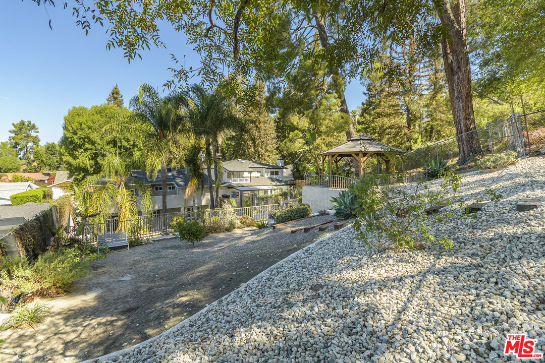 House set among mature trees and palms, with a rocky hillside garden and covered gazebo.